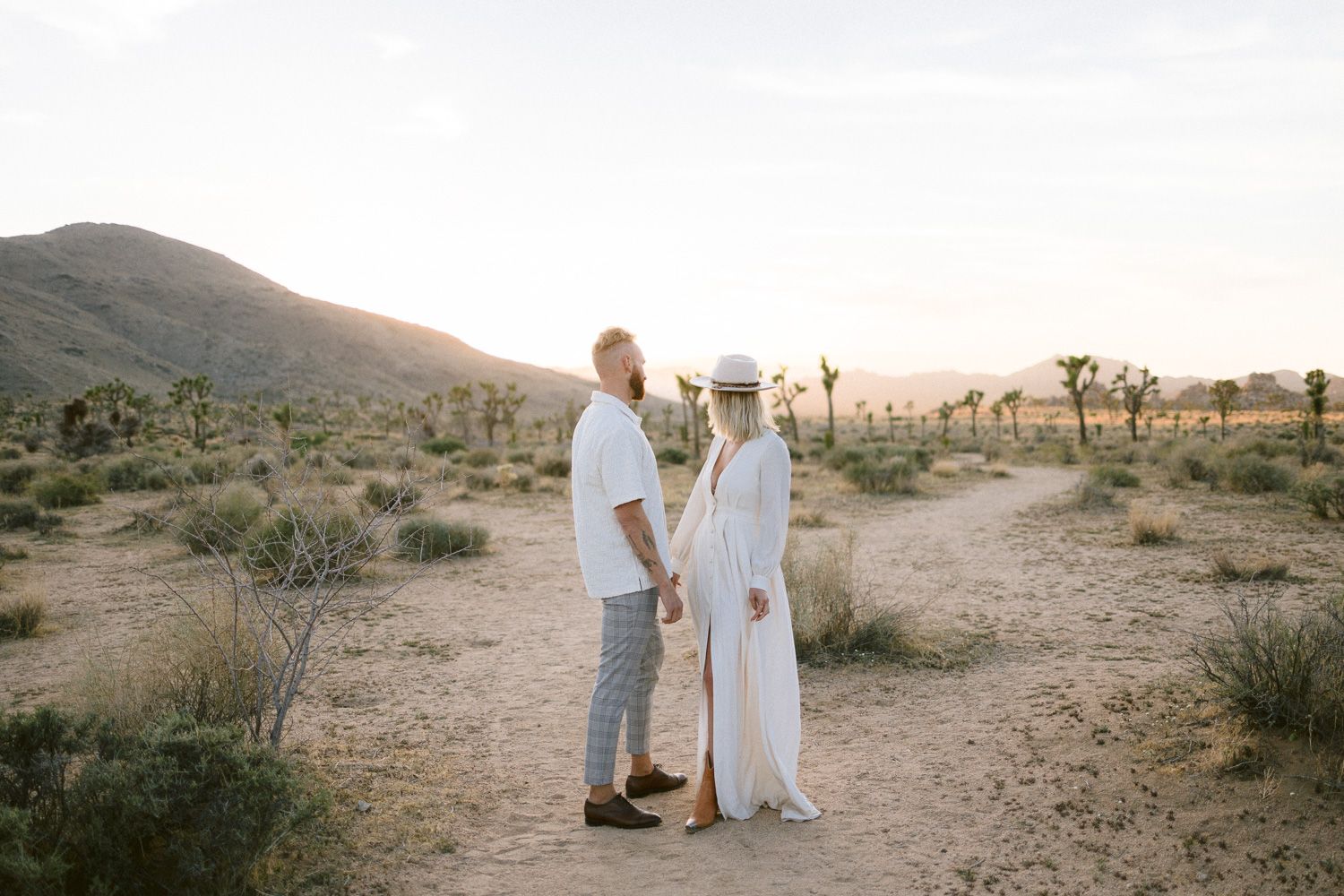 A couple stands together on a desert path with mountains and Joshua trees at golden hour.