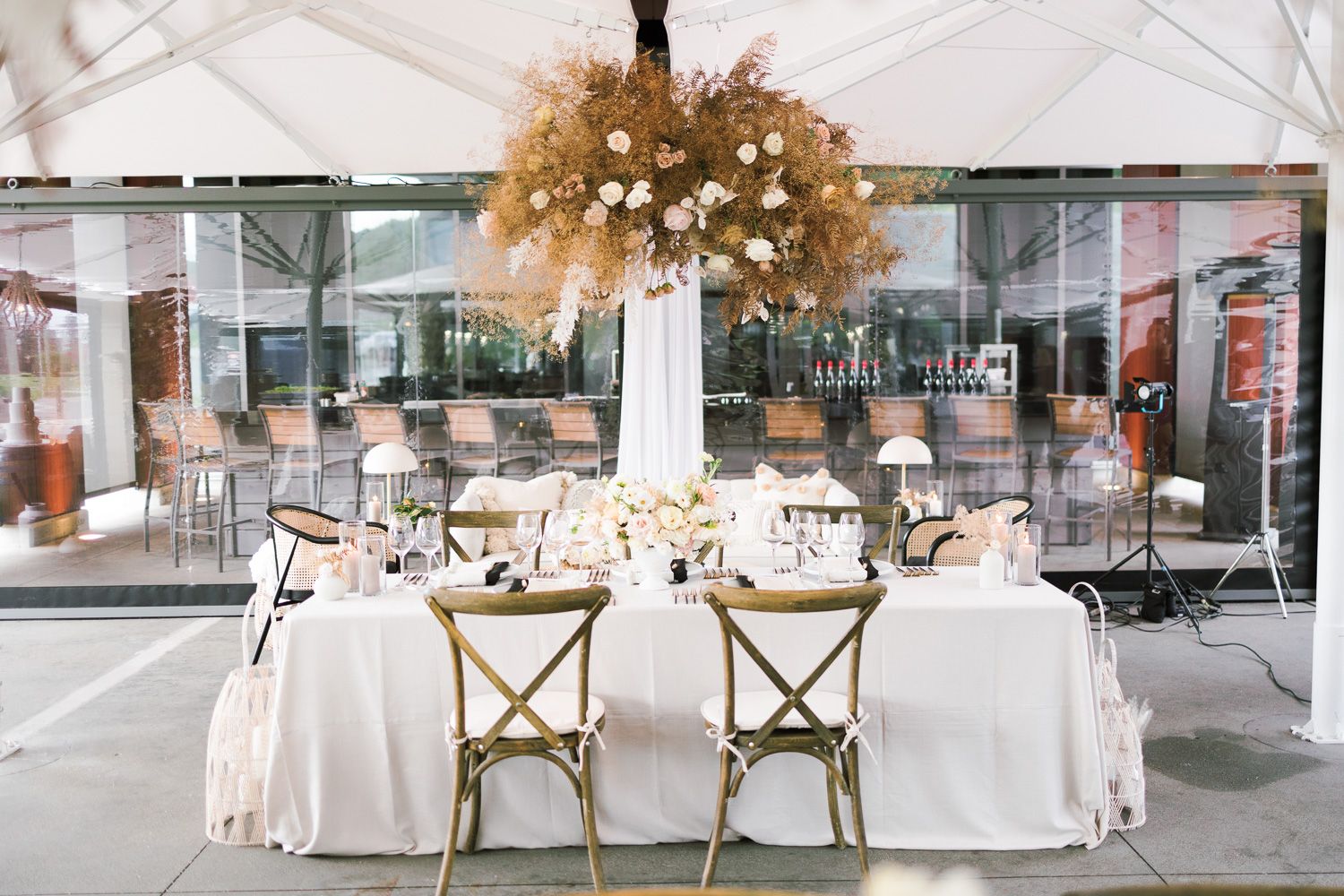 Outdoor dining setup with rustic crossback chairs and dramatic dried floral centerpiece under a white tent.