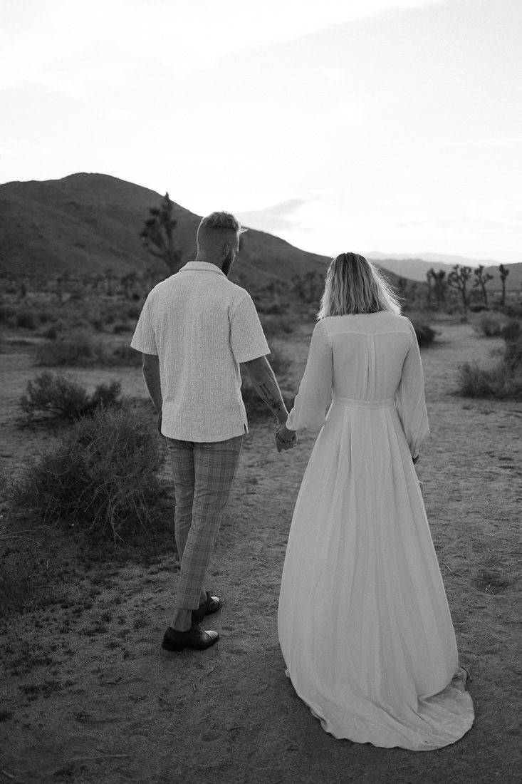 Black and white photo of couple walking through desert with Joshua trees and mountains.