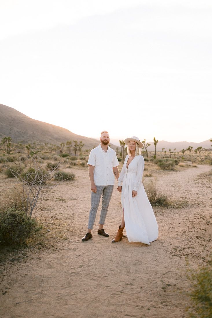 Desert engagement photo featuring a couple in white and plaid standing among Joshua Trees at sunset.