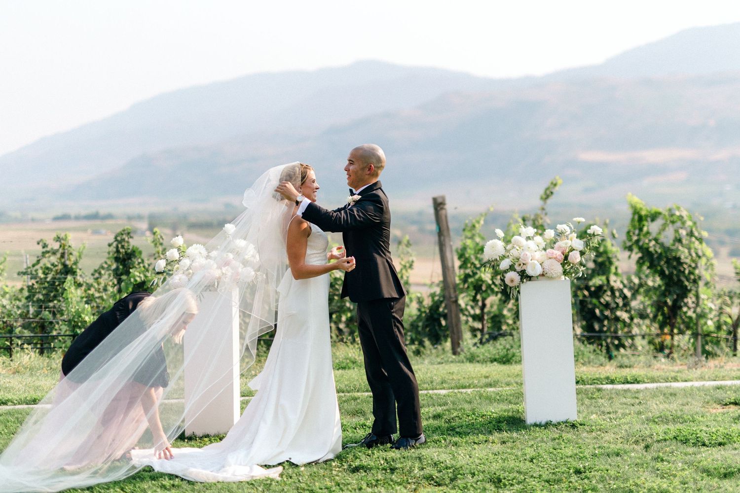 A couple celebrates their wedding ceremony in a scenic vineyard setting with mountains in the background.