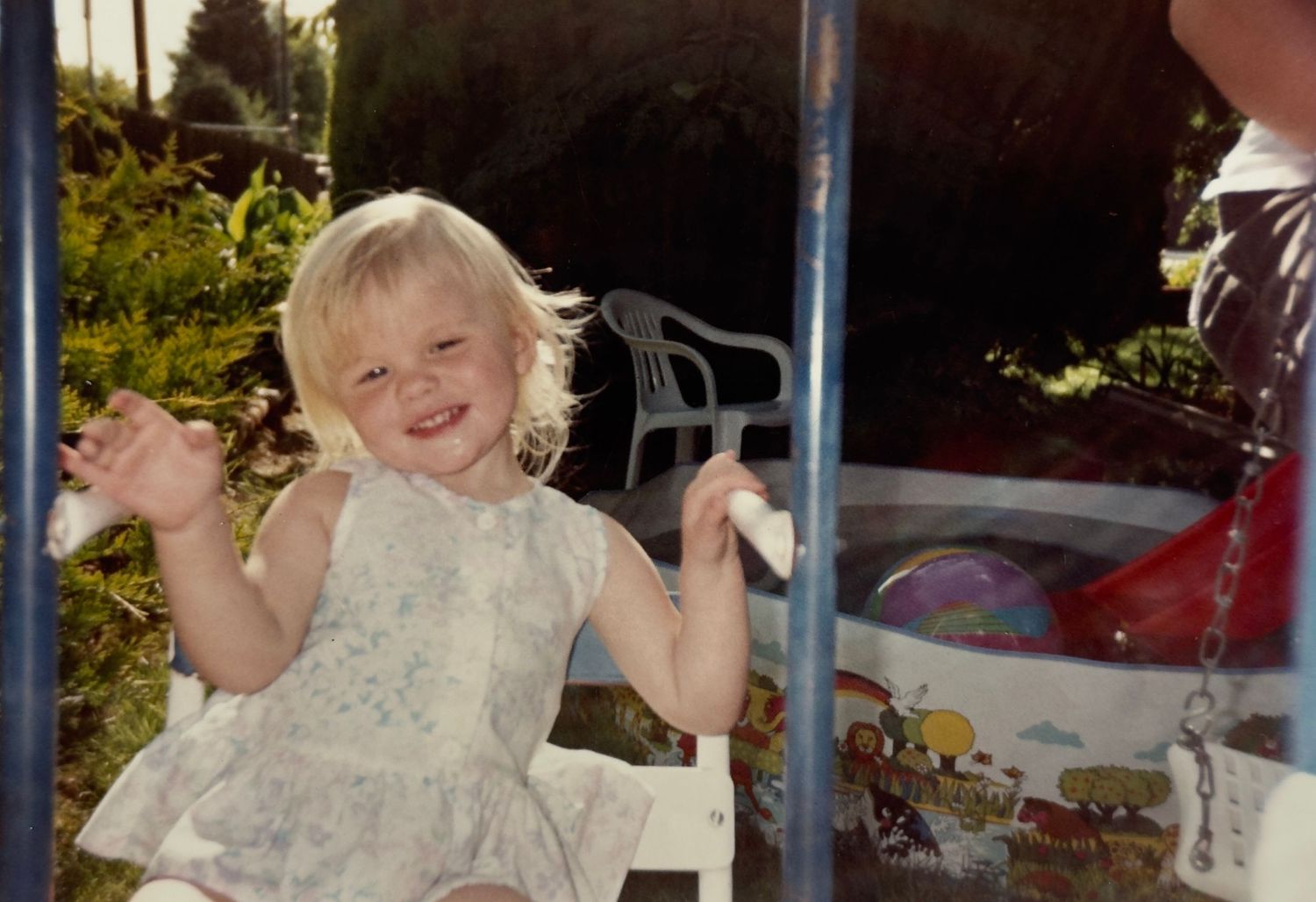 A young child in a white dress laughs joyfully while swinging on outdoor playground equipment in a sunny garden setting.