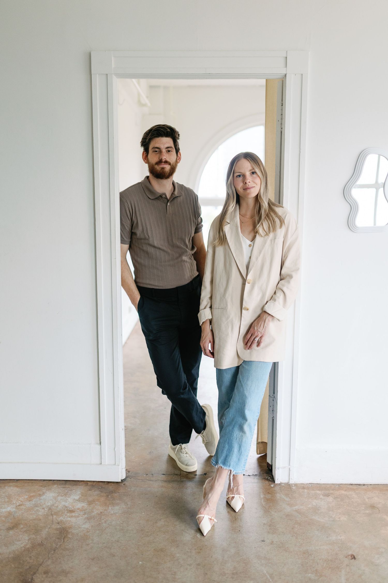 Couple poses casually in doorway wearing trendy minimalist outfits with brown sweater and beige blazer with jeans.