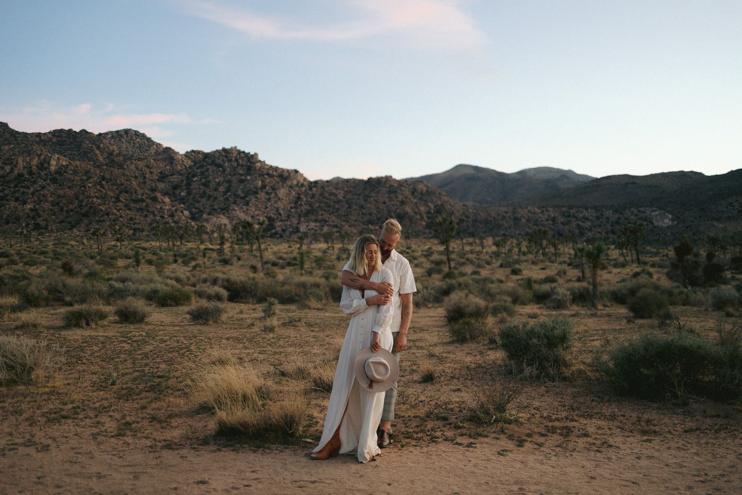 Two people embrace while standing in a desert landscape during golden hour.