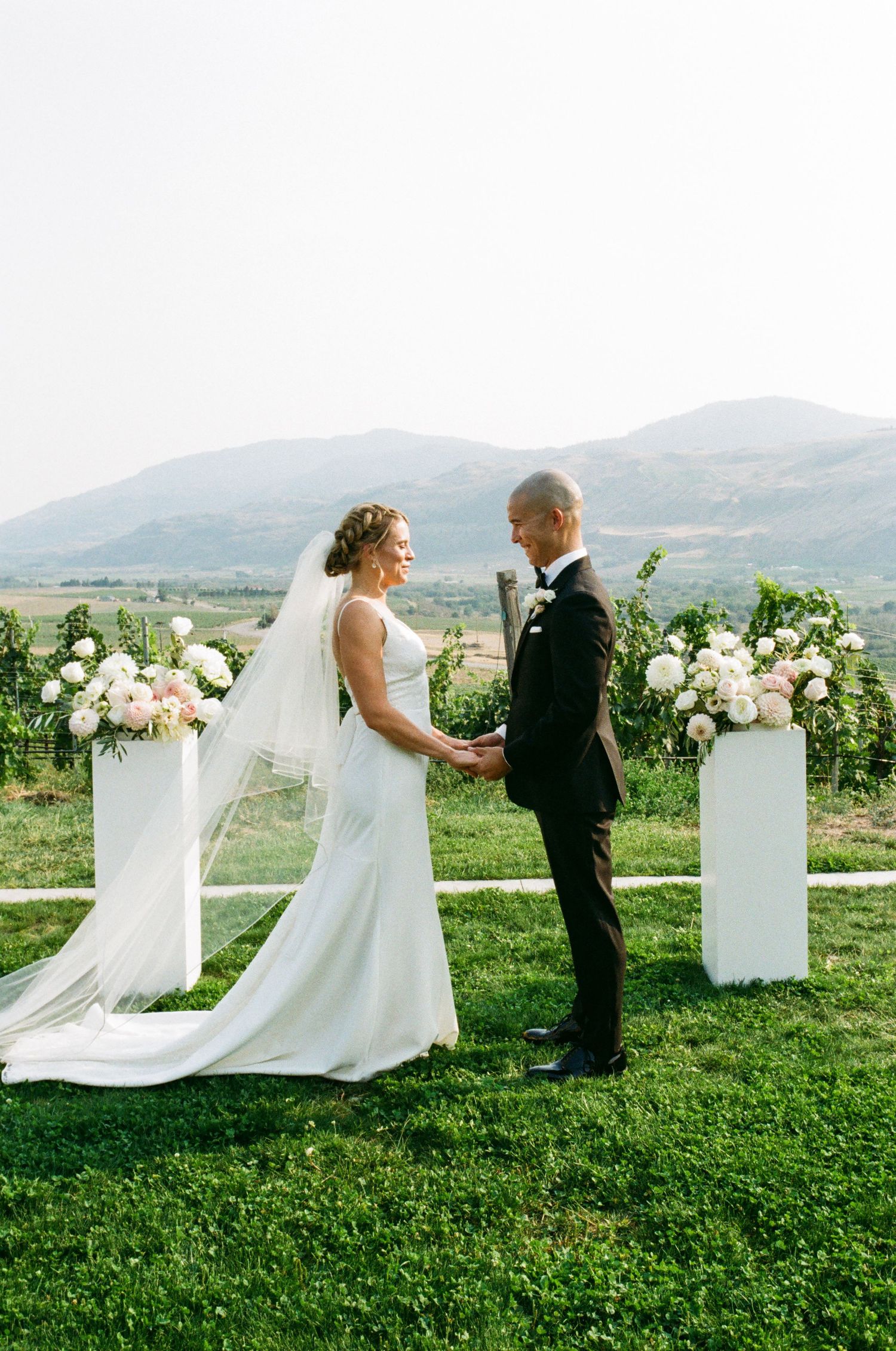 Wedding couple poses for portraits on a grass lawn with mountain scenery and white pedestals.