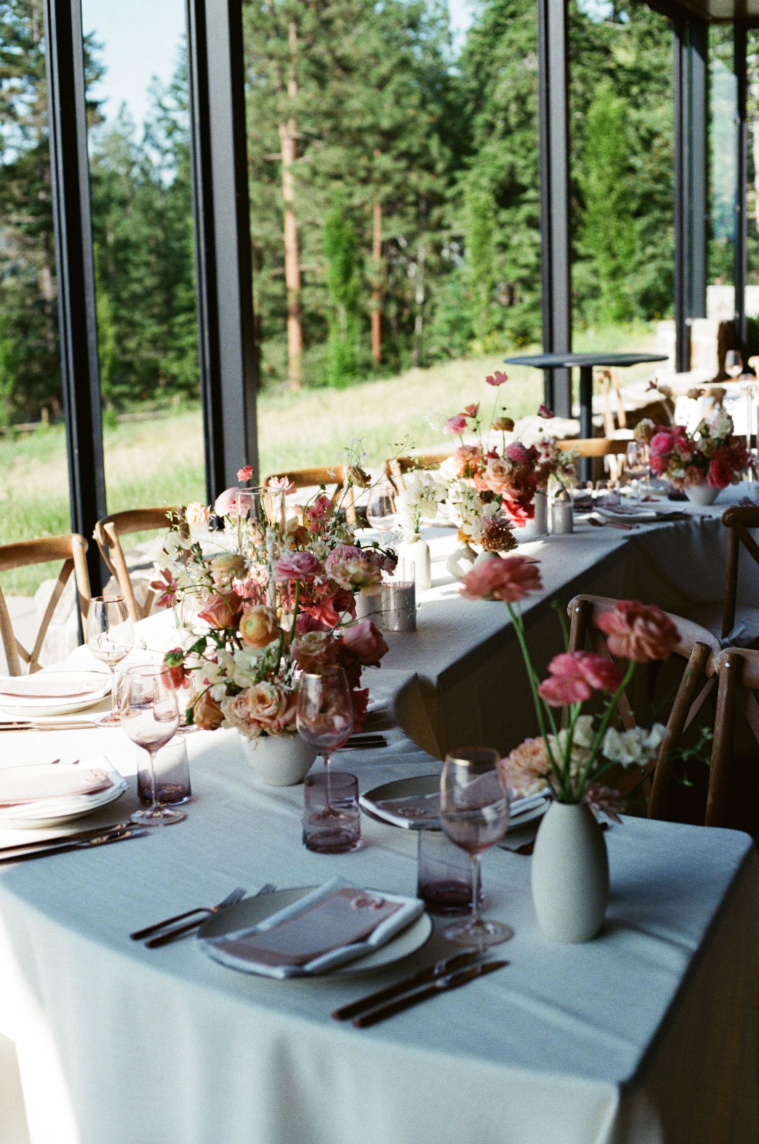 Elegant table setting in modern restaurant with white linens, pink floral arrangements and forest views through large windows.