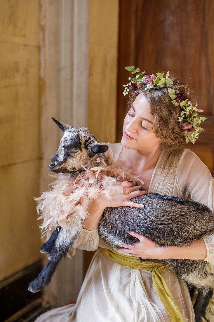 A dreamy portrait featuring someone in a floral crown holding a baby goat while wearing a romantic cream-colored dress.