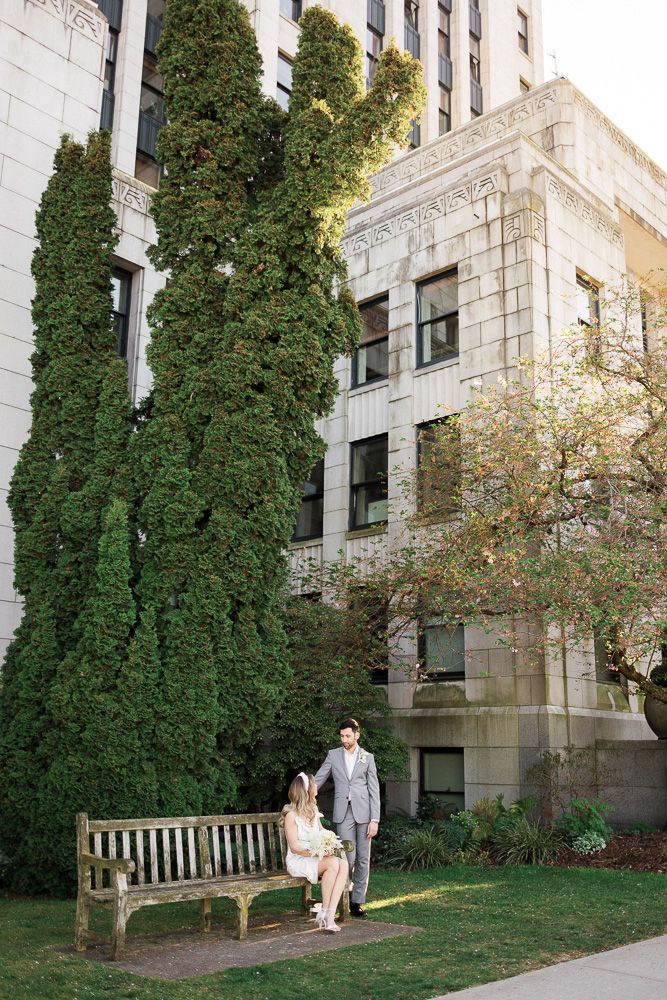 Wedding couple seated on wooden bench against classic stone building with tall evergreens.