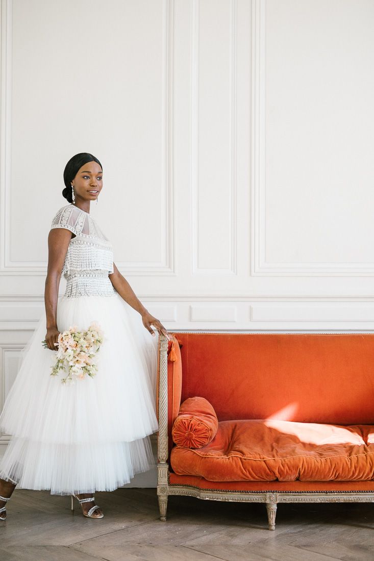 Bride in tea-length tulle dress stands beside orange vintage sofa in elegant white room.