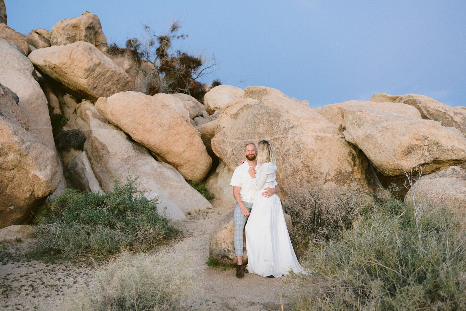 Couple in white clothing standing together among large desert boulders under blue sky.