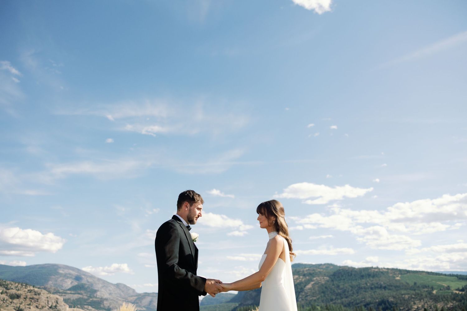 A couple holds hands on a mountaintop with blue skies and rolling hills in the background.