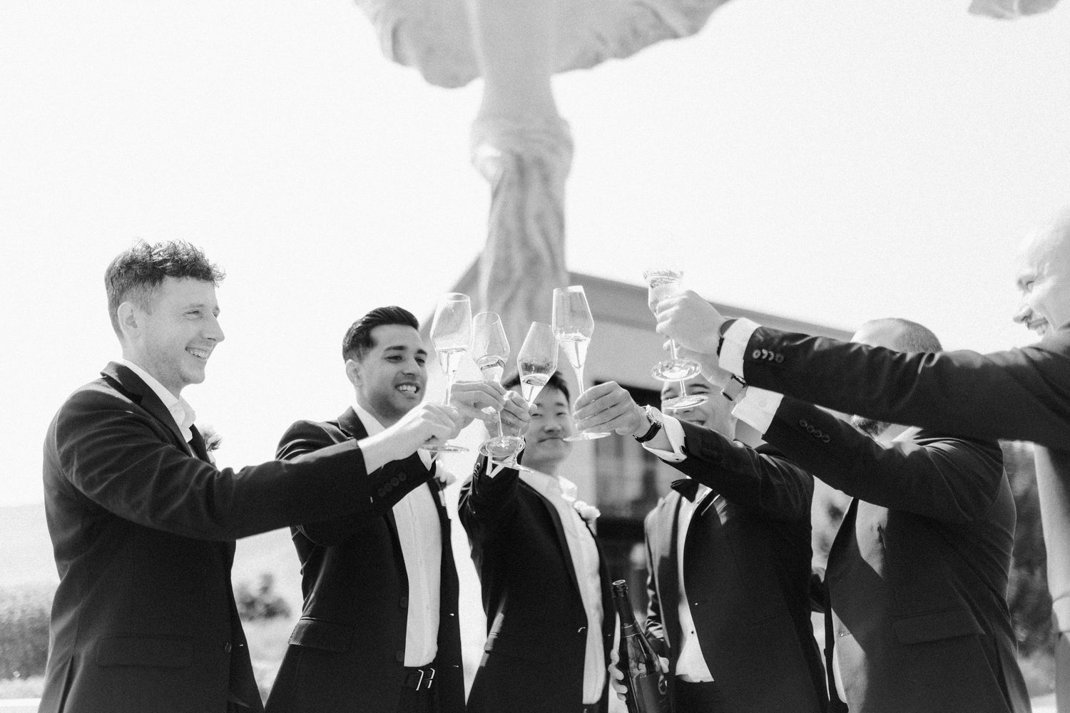 A group of groomsmen in black suits perform a coordinated jumping pose outdoors during a wedding photo session.