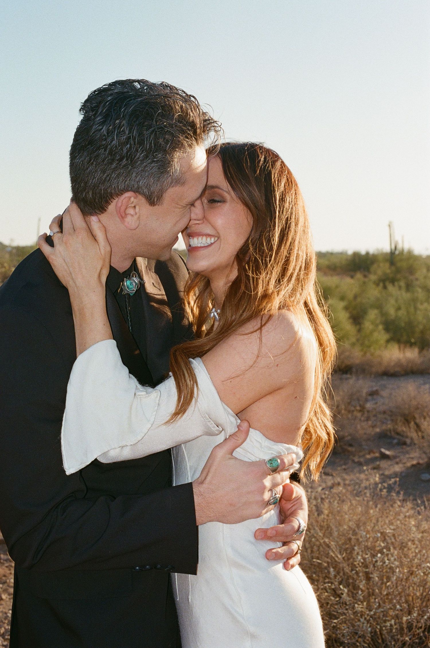 A romantic desert wedding photo of a couple embracing and smiling at sunset with cacti in the background.