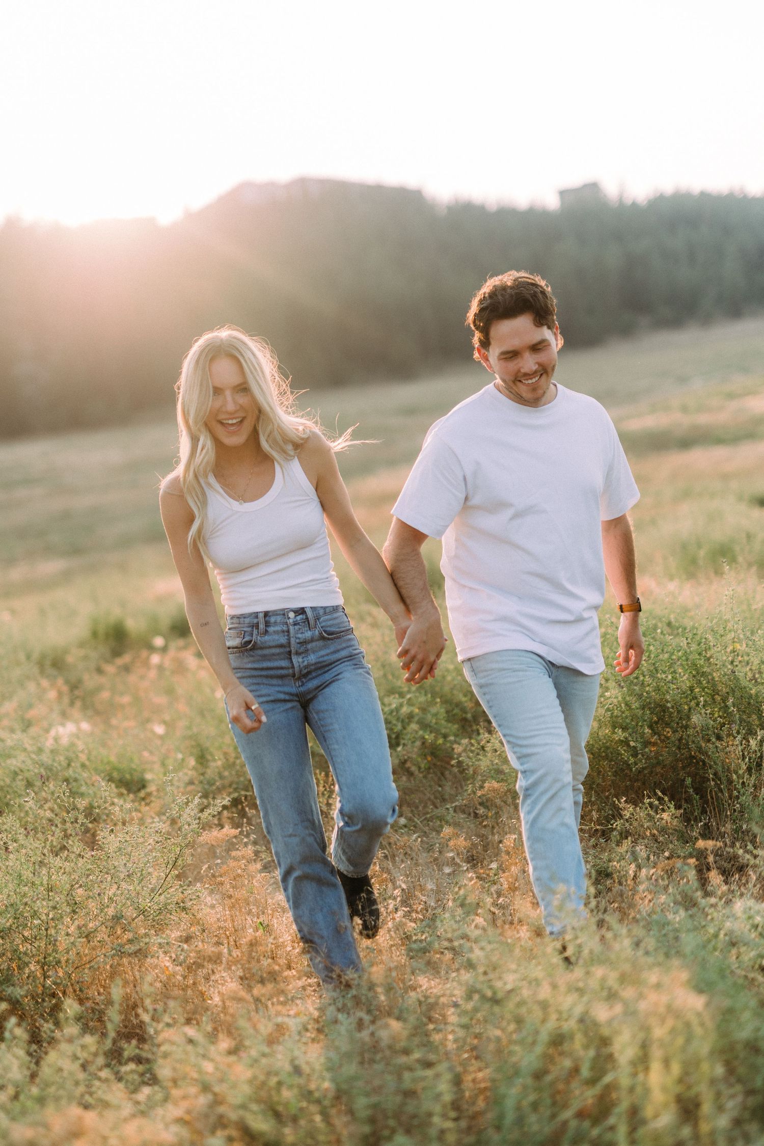 Couple walking hand in hand through a golden field at sunset in casual summer attire.