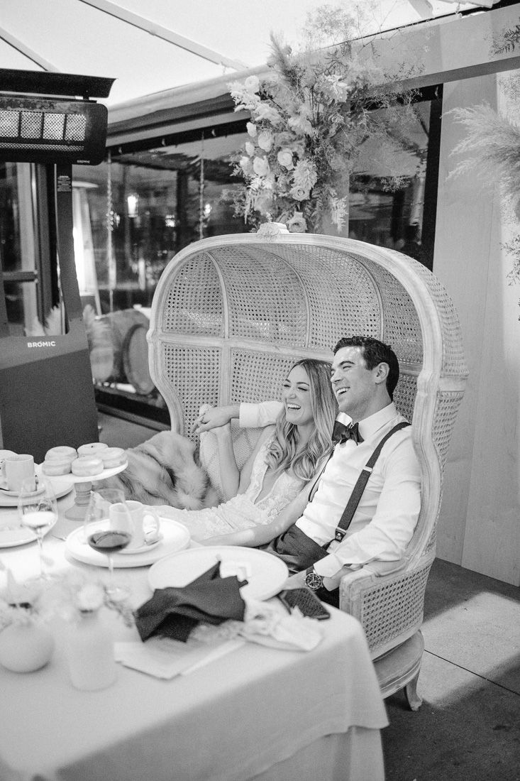 Couple shares joyful moment while seated in peacock chair during wedding reception.
