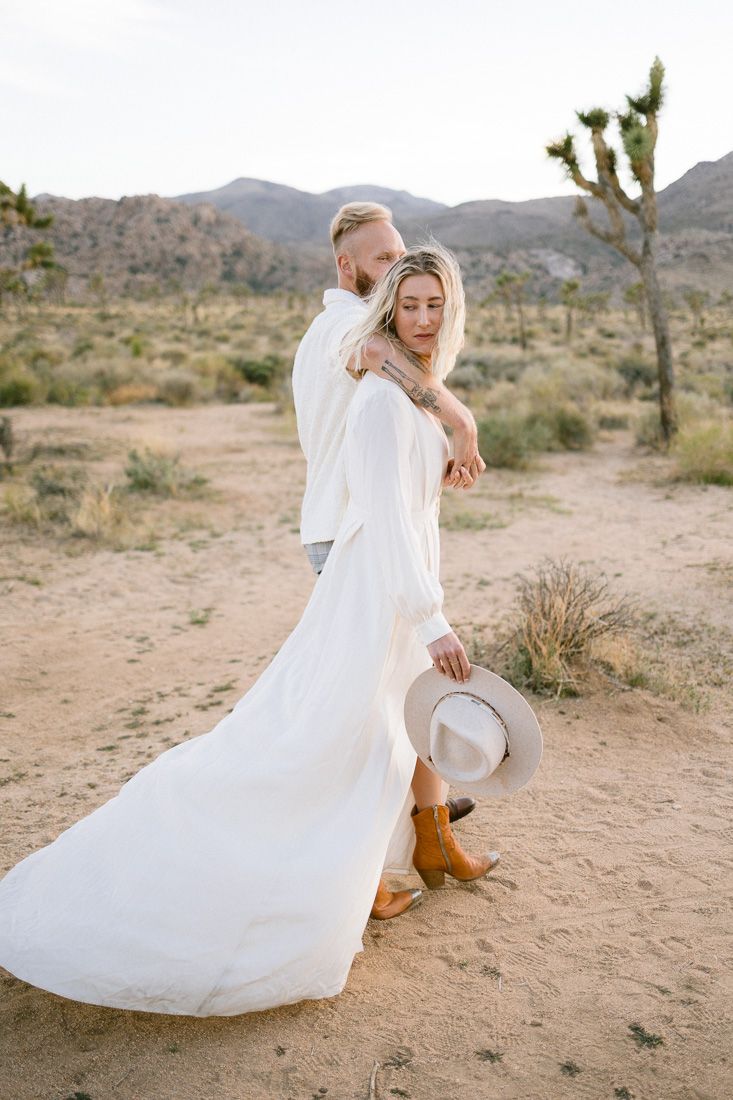 Couple in white embracing in a desert landscape with mountains and Joshua trees in the background.