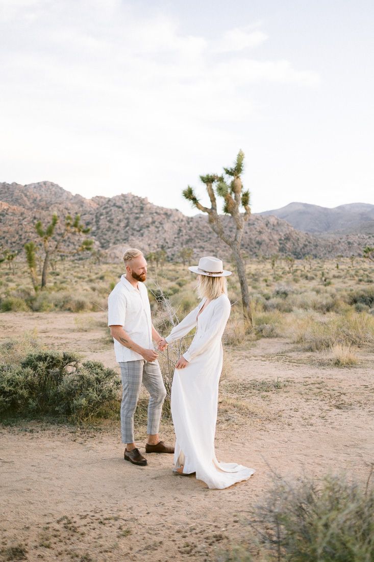Couple in light colored clothing standing together on a desert trail with Joshua trees and mountains.