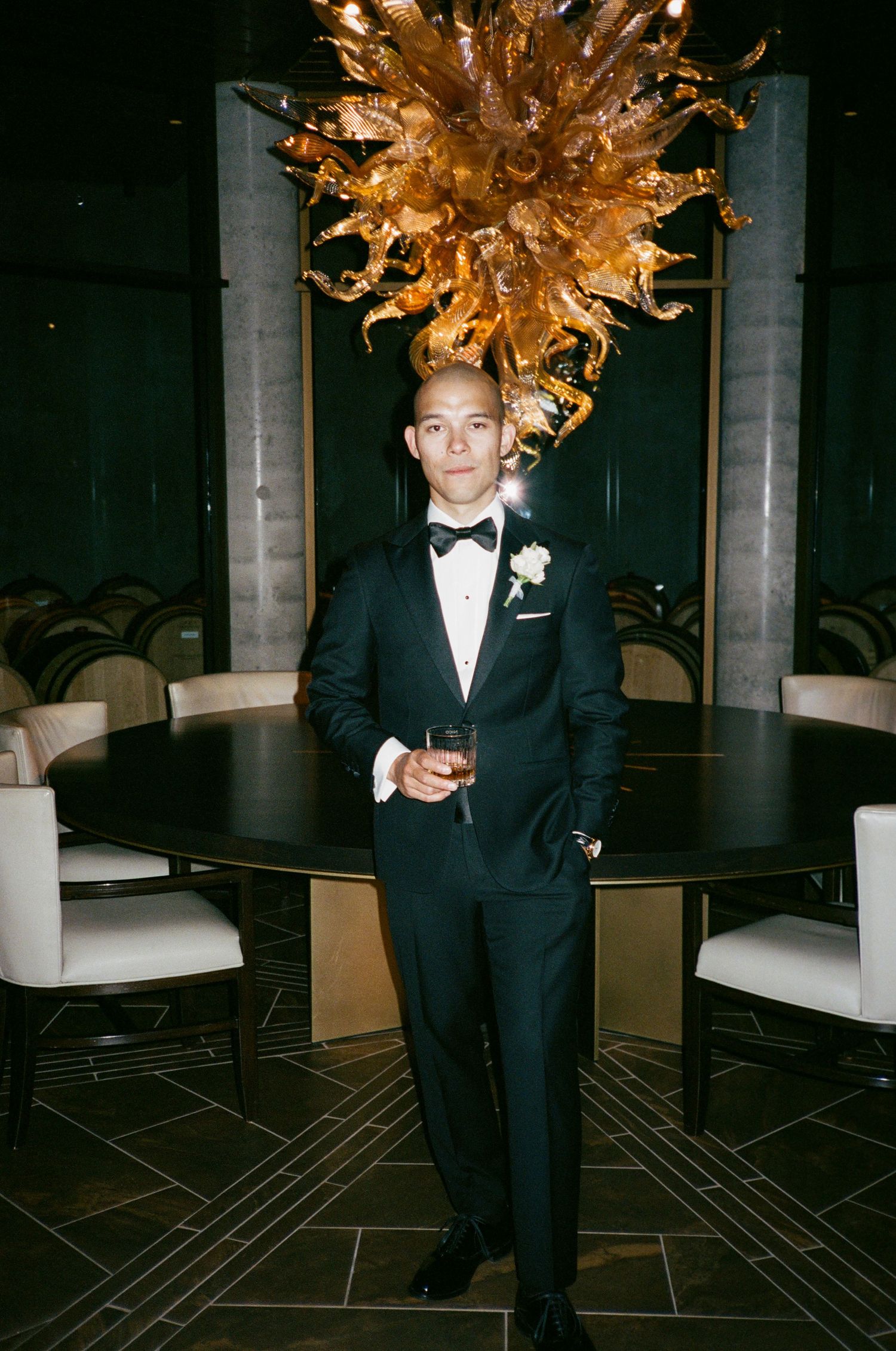 Elegant man in tuxedo poses beneath dramatic gold chandelier in formal dining setting.