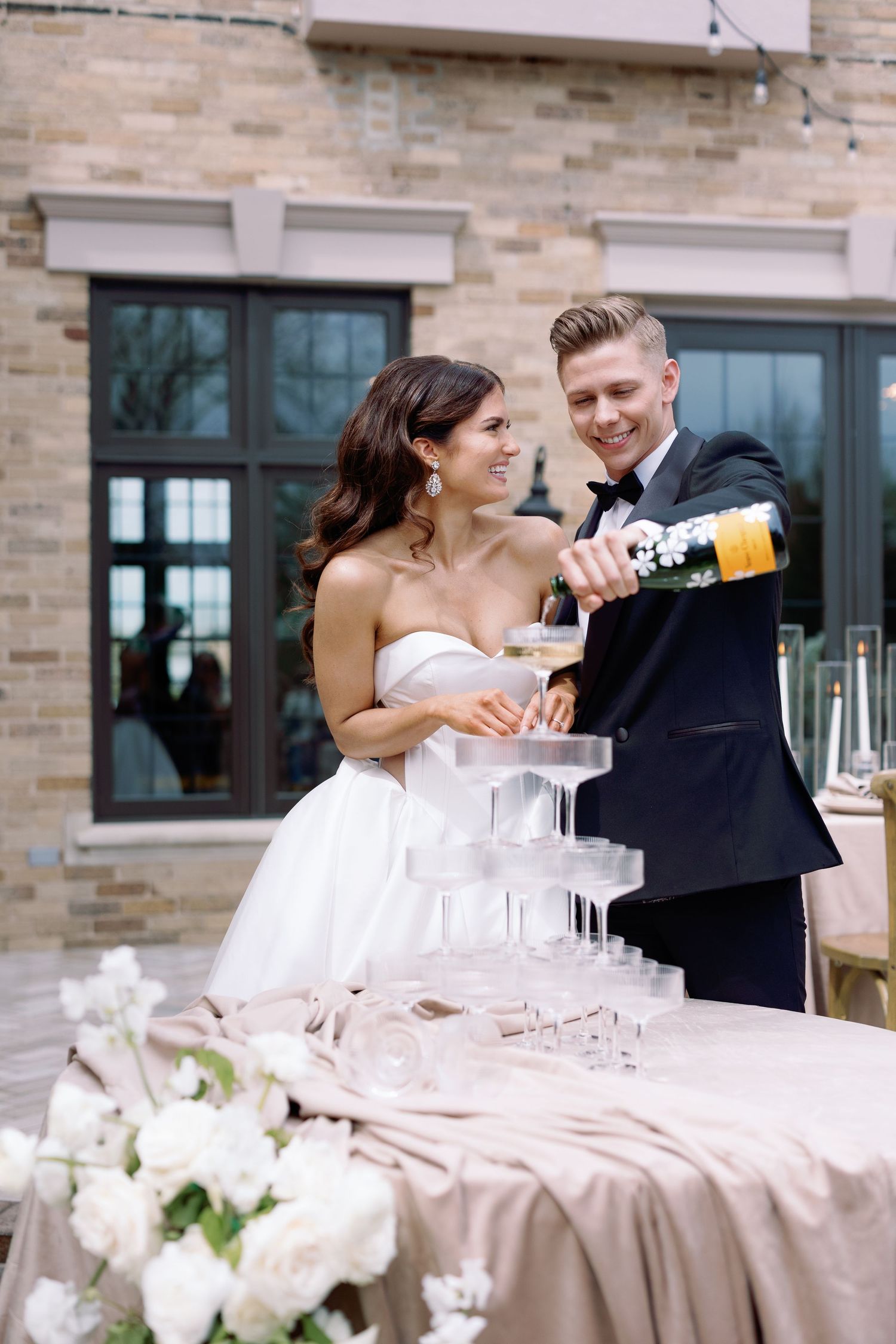 A couple pours champagne into a tower of glasses at an elegant wedding reception with white floral decorations.