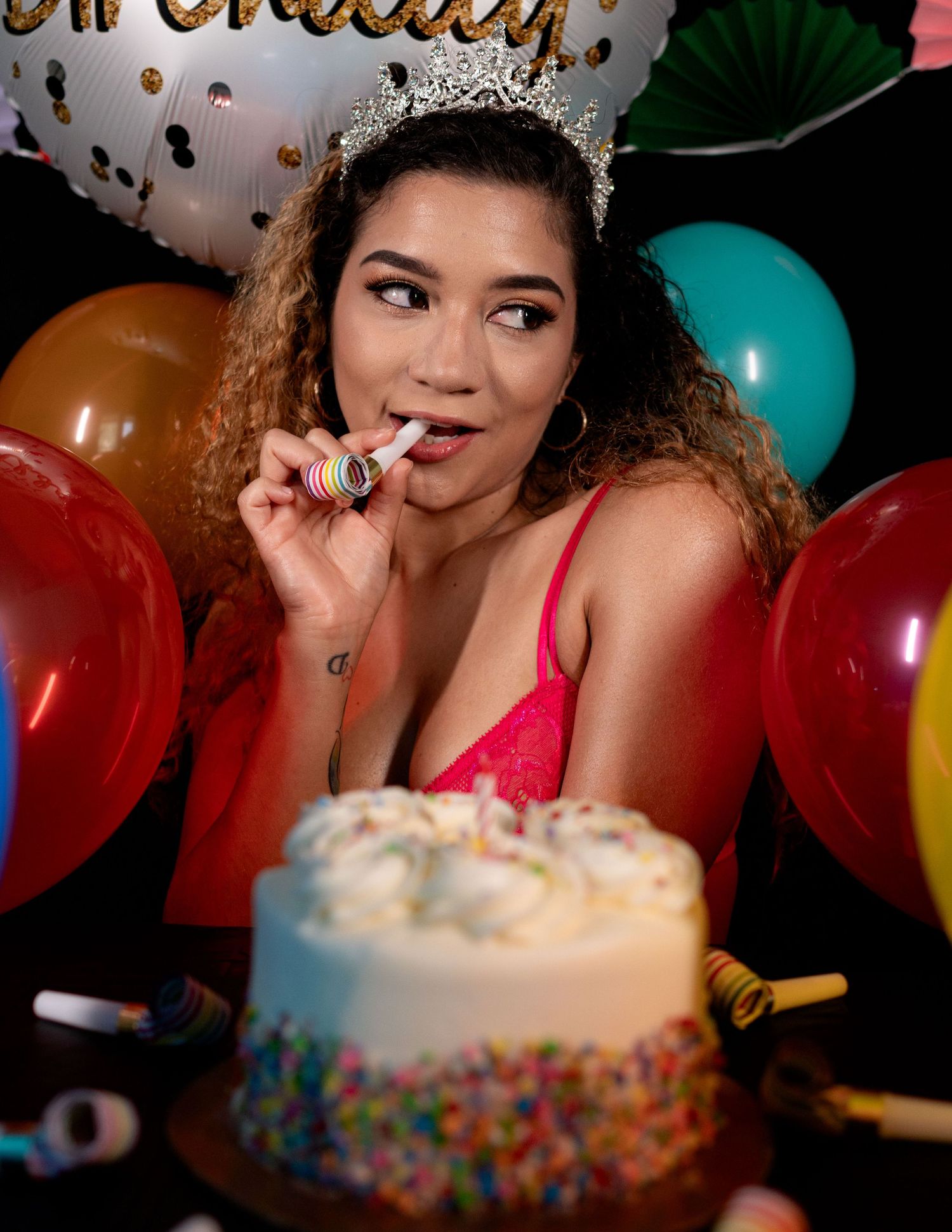 Woman in tiara and pink lace top celebrates birthday with cake, balloons, and party blower.