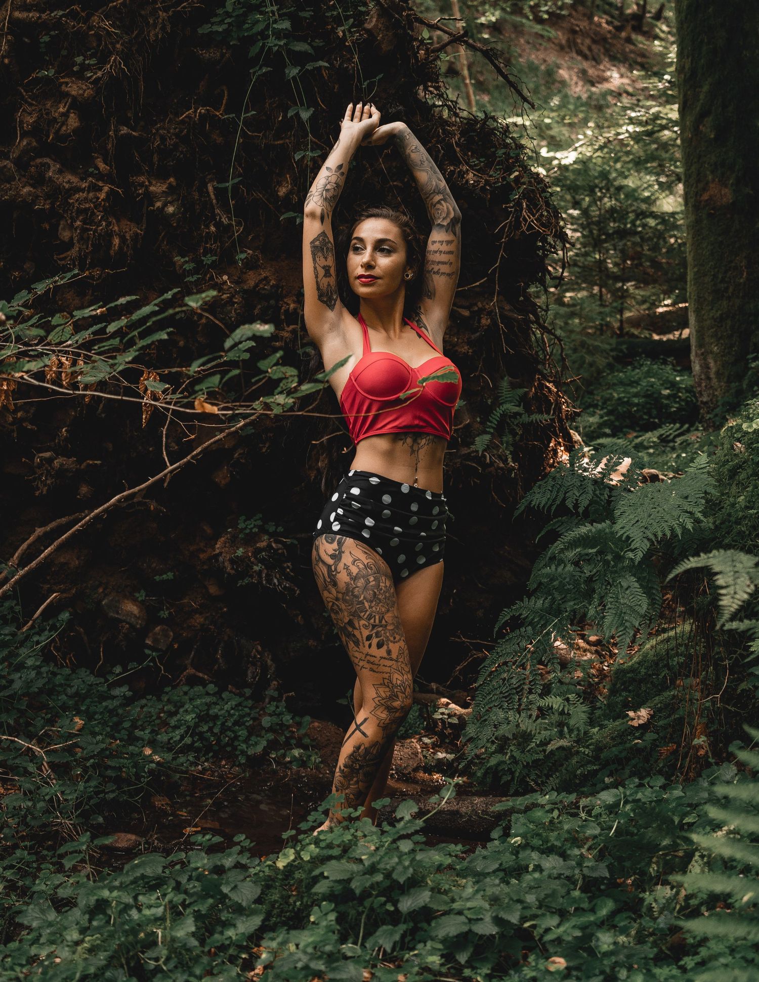 Tattooed woman in red bikini top and polka dot shorts posing in lush green forest setting.