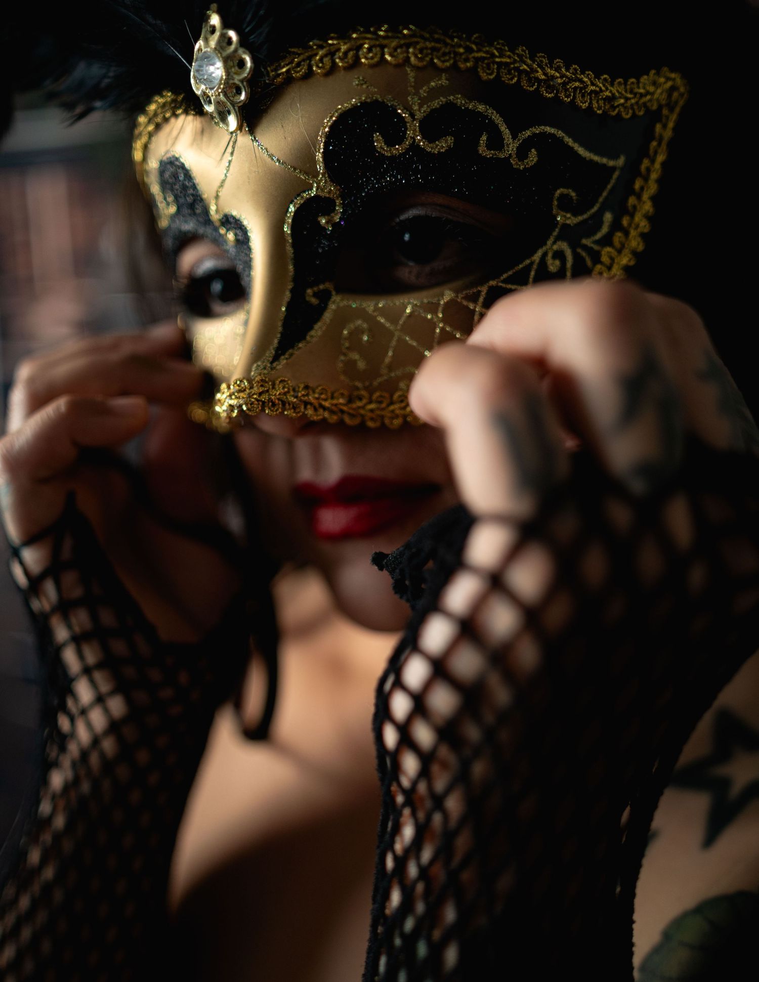 Woman in gold and black Venetian masquerade mask with fishnet gloves in dramatic dark setting.