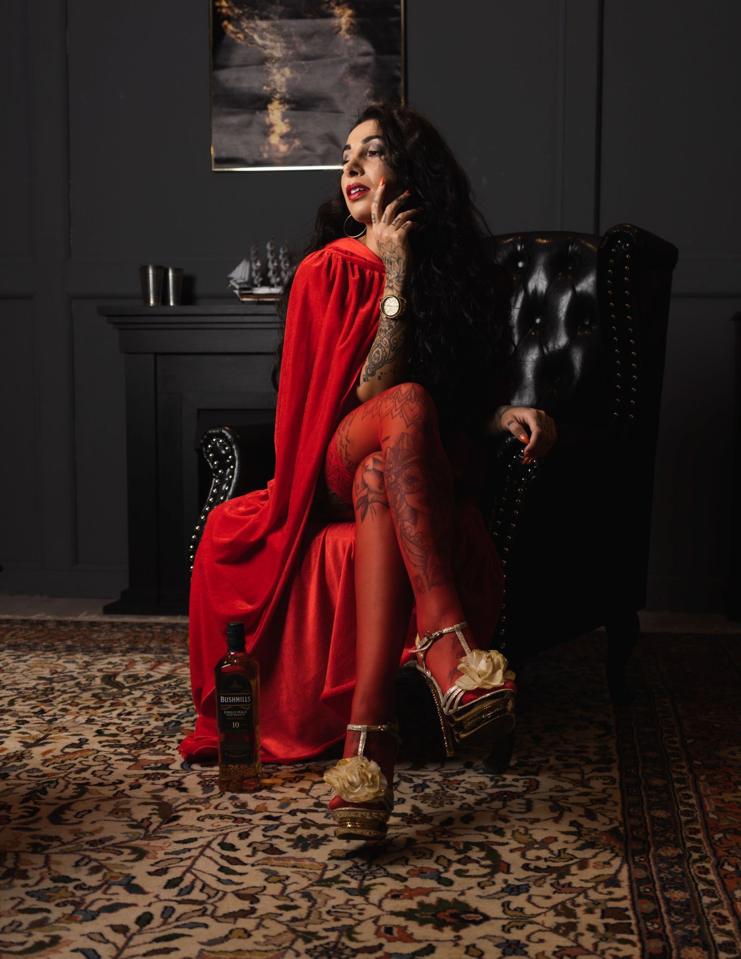 Tattooed woman in red dress and cape sits in leather chair, smoking, with a bottle nearby in dark elegant room.