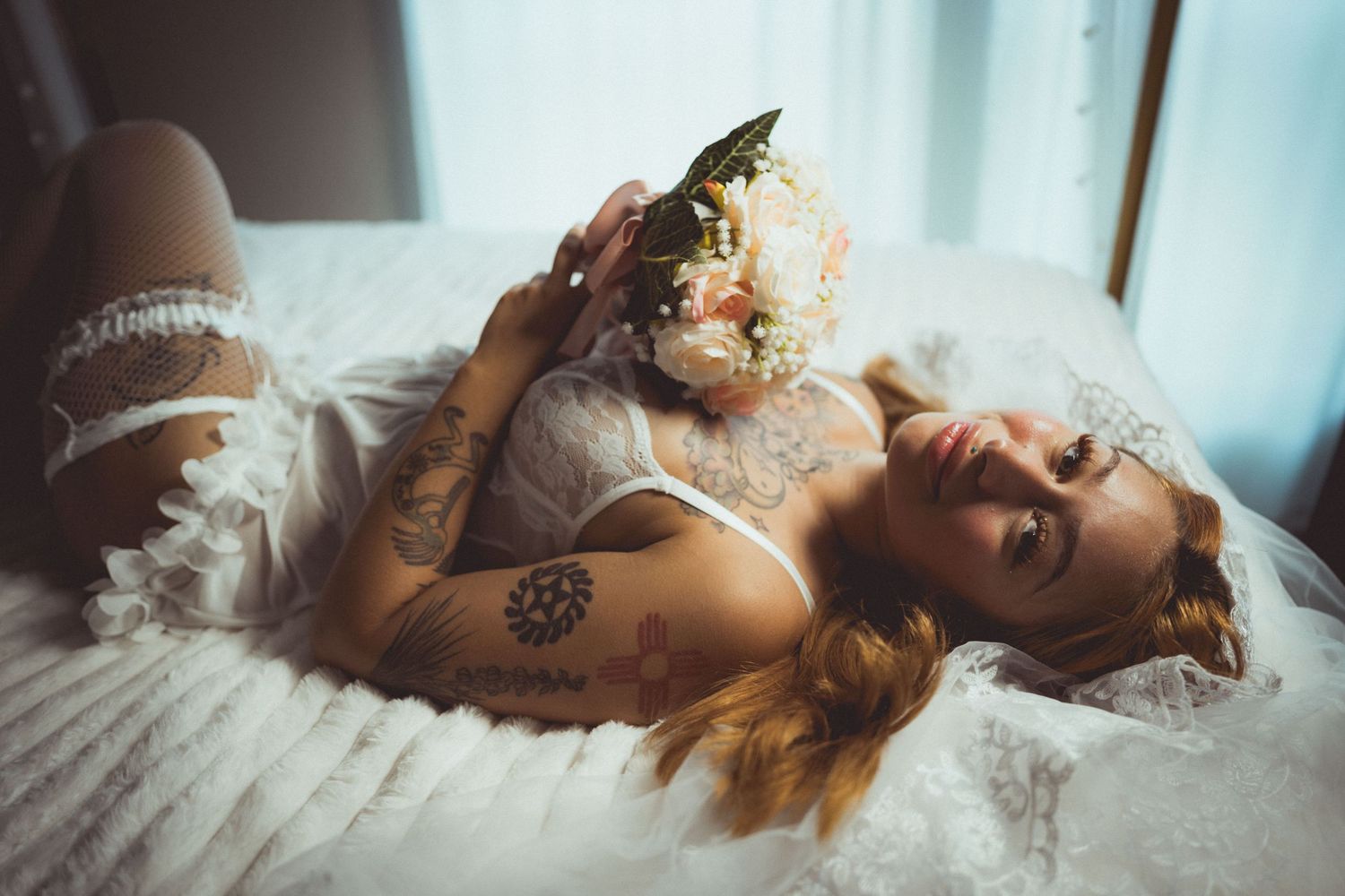Tattooed bride in lace lingerie poses with floral bouquet on white bed.