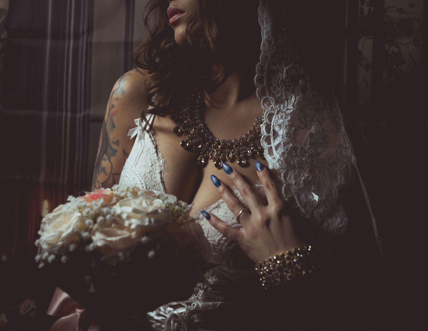 Tattooed bride in lace dress holding floral bouquet, wearing ornate necklace and blue nail polish with rings.