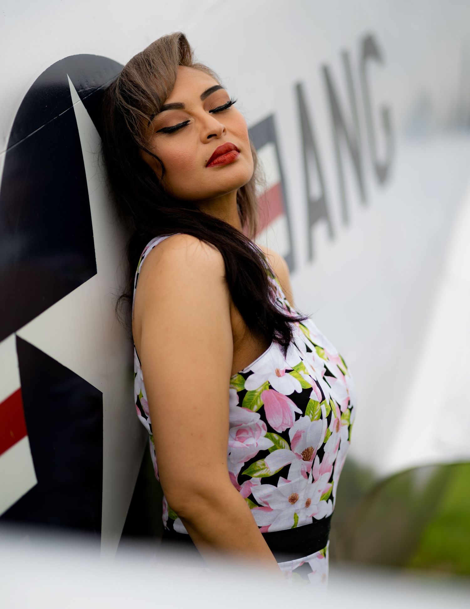 A person in a floral print dress poses against a geometric backdrop with closed eyes and red lipstick.