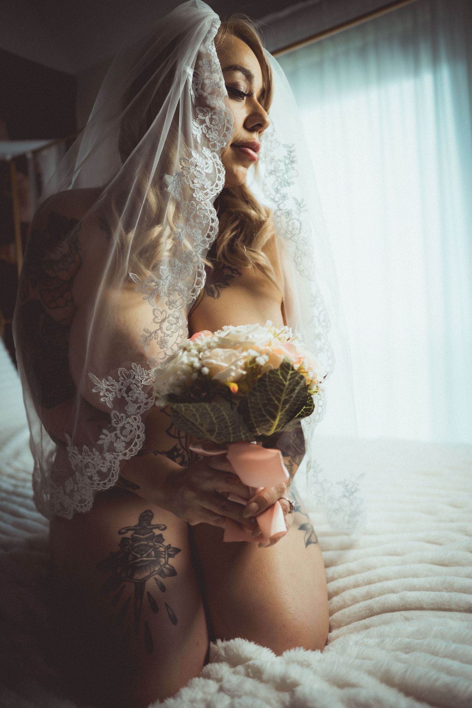 Tattooed bride in lingerie and lace veil kneeling on bed holding bridal bouquet in soft natural light.