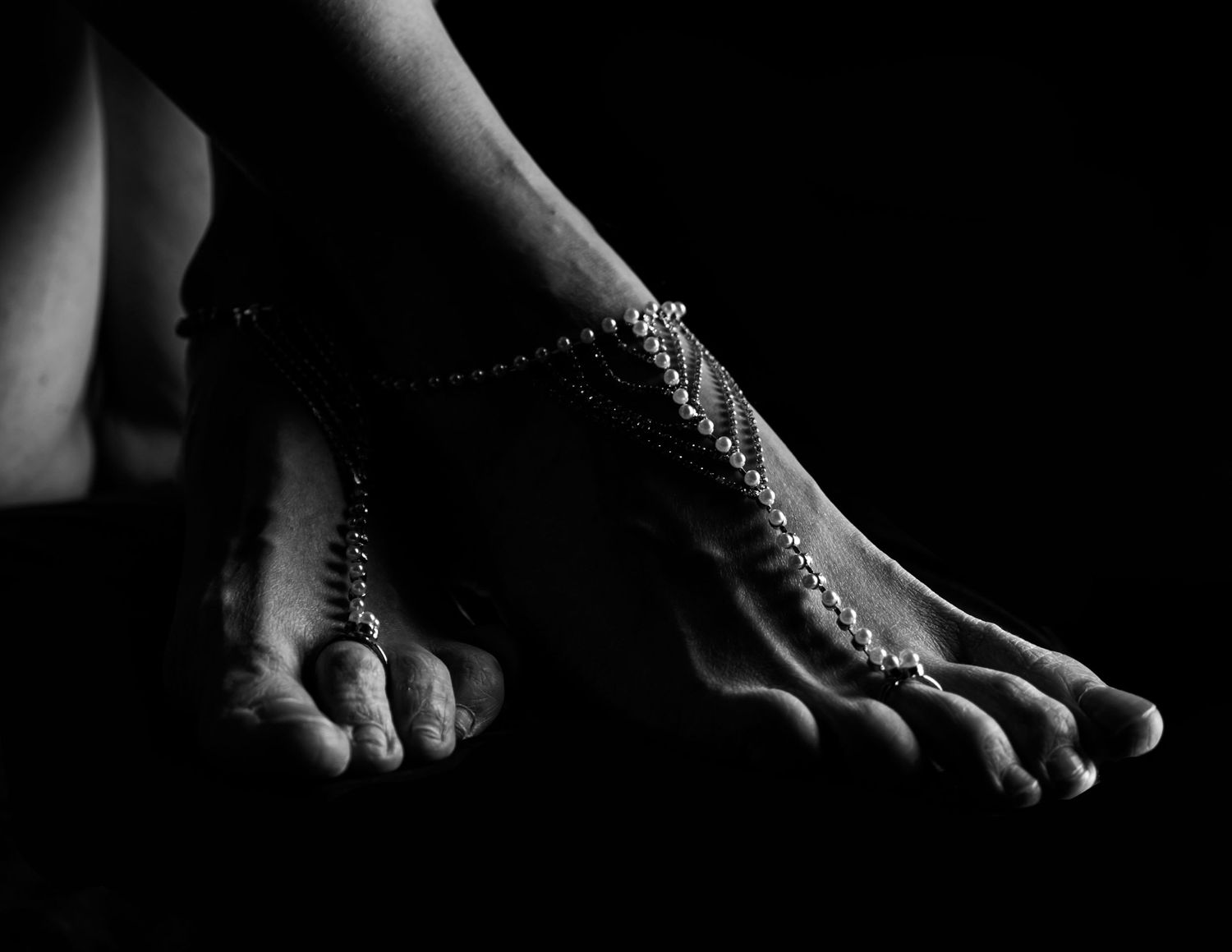 Black and white artistic photograph of bare feet with delicate jewelry chains draped across them in dramatic lighting.