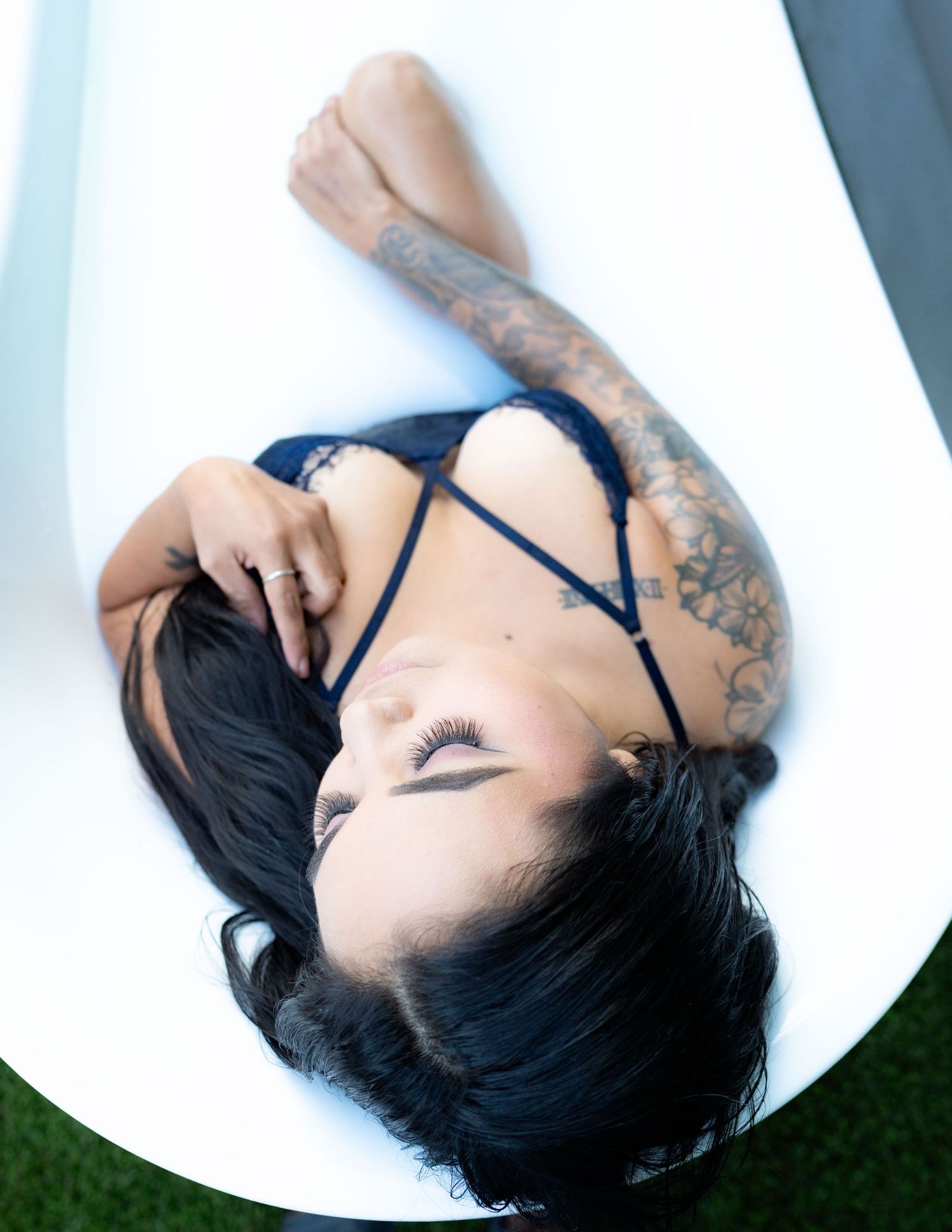 Woman with tattoos lying in a white bathtub wearing black lingerie, eyes closed, dark hair spread out.