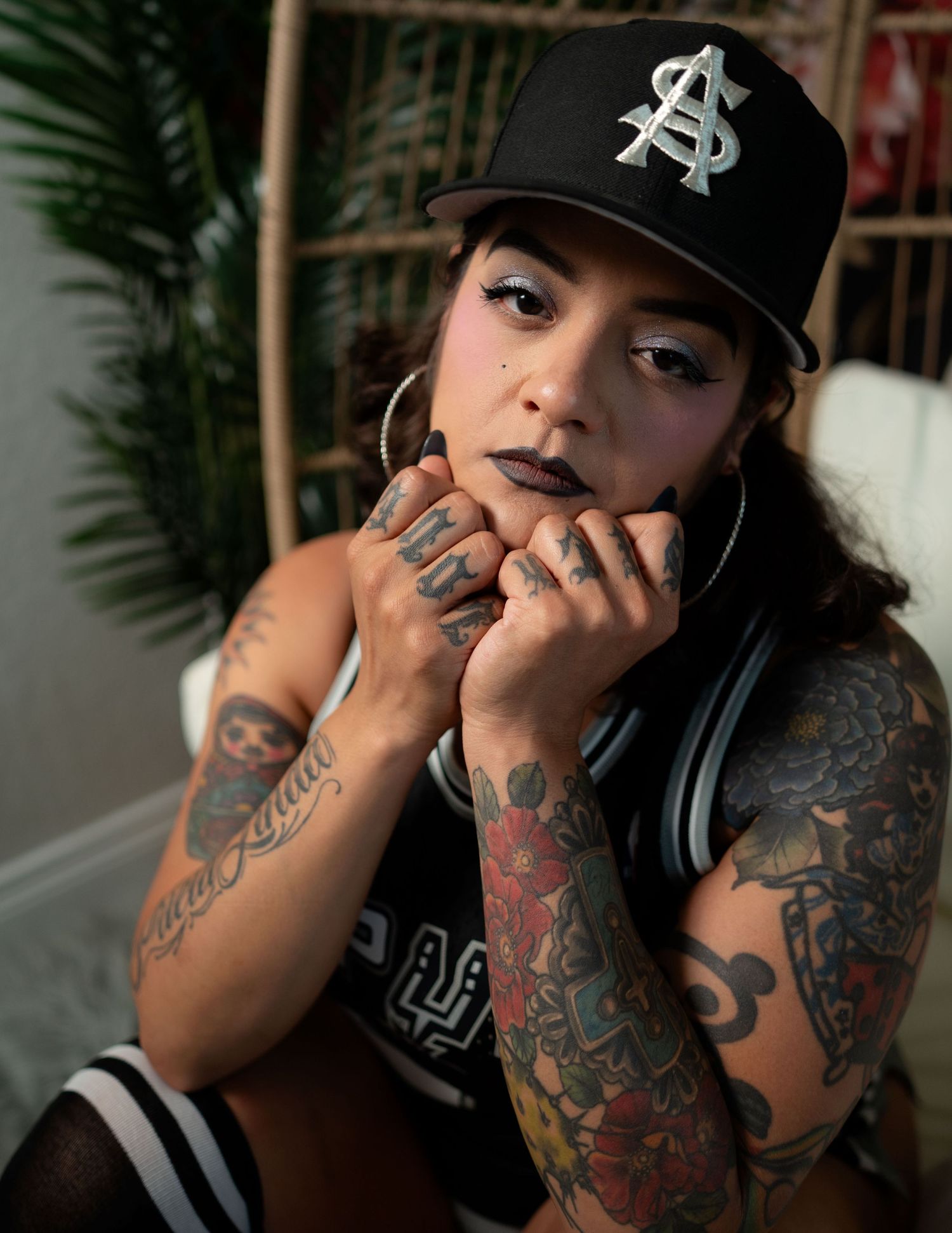 Tattooed woman wearing a black snapback cap and hoop earrings, posing with hands near face.
