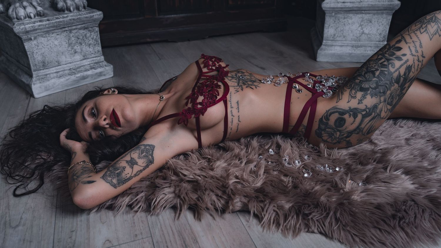 Tattooed woman in red lingerie posing on fur rug in elegant room with ornate decor.