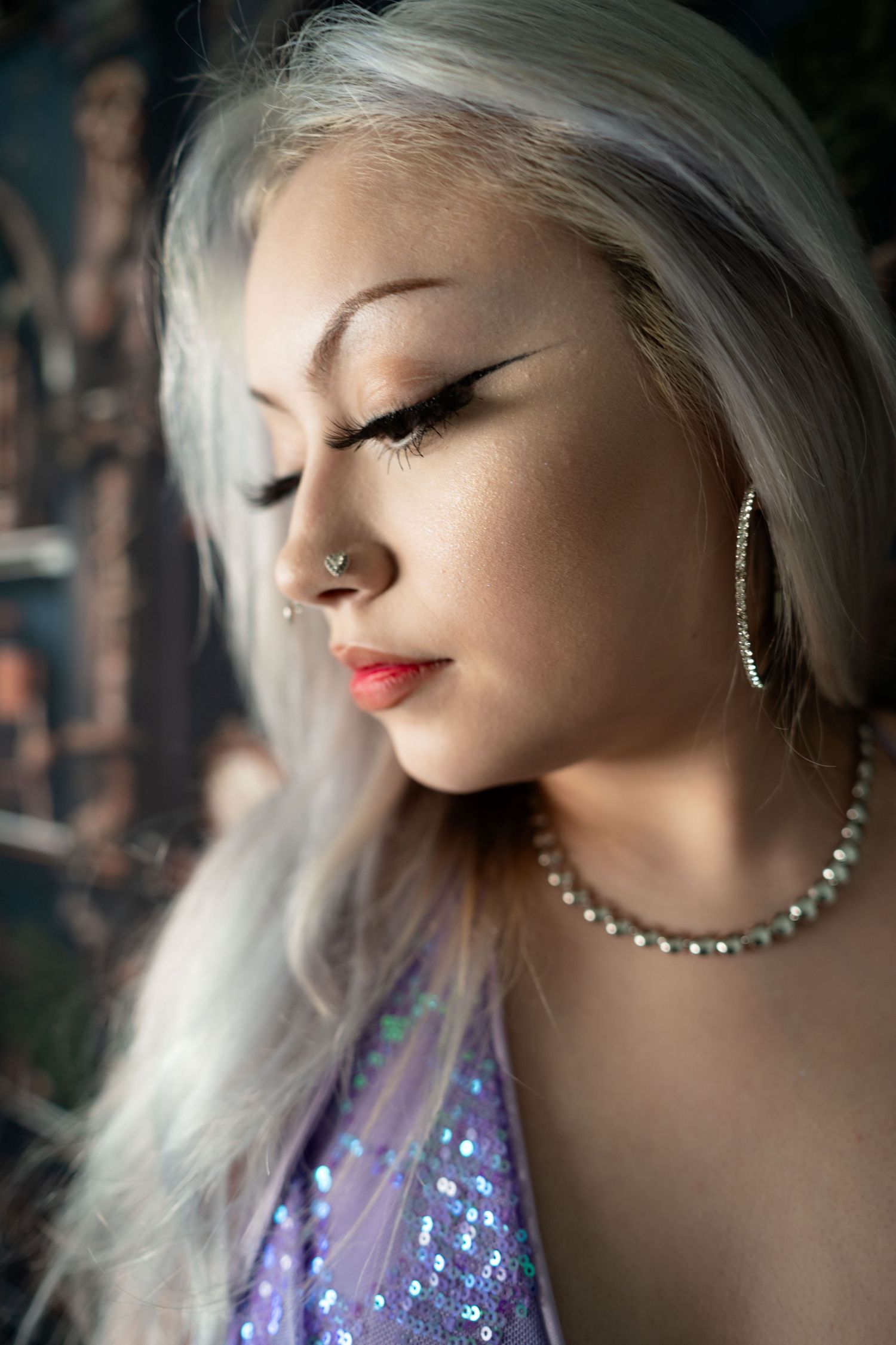 Young woman with silver hair, bold eyeliner, nose ring, pearl necklace, and sequined lavender top poses with eyes closed.