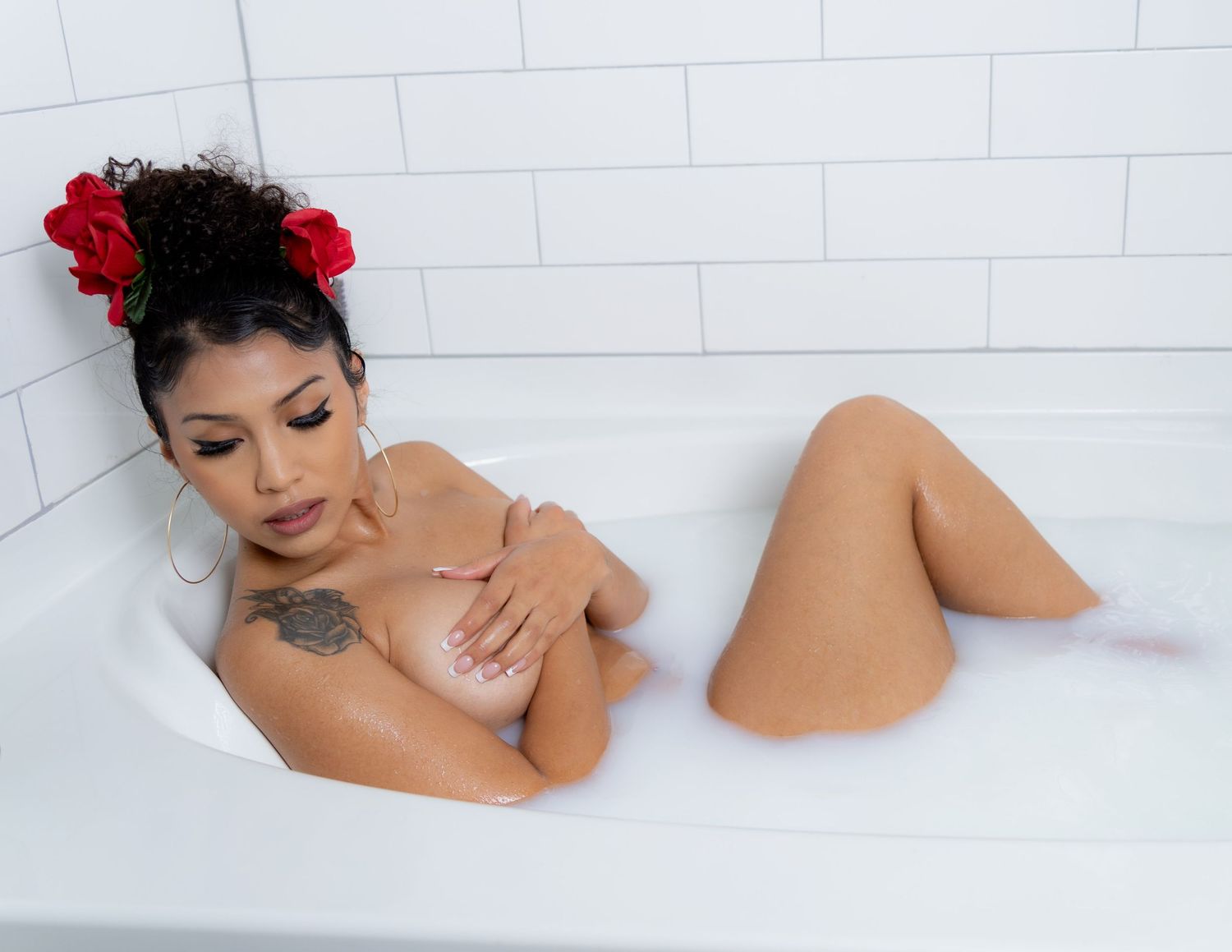 Woman with red roses in hair relaxing in a bubble bath, showcasing a shoulder tattoo.
