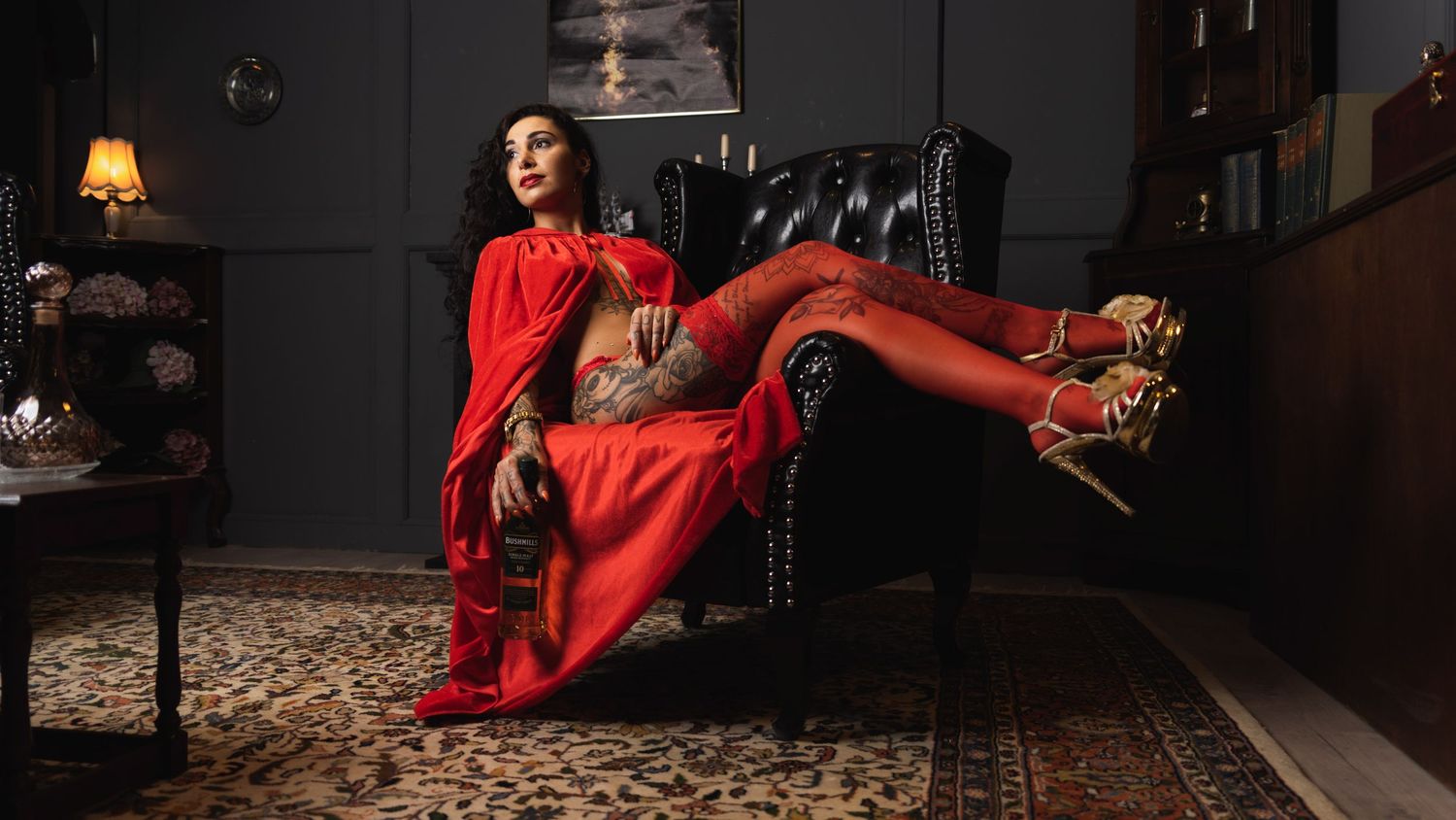 A figure in a flowing red dress and heels lounges elegantly across a vintage leather armchair in a moody dark room.