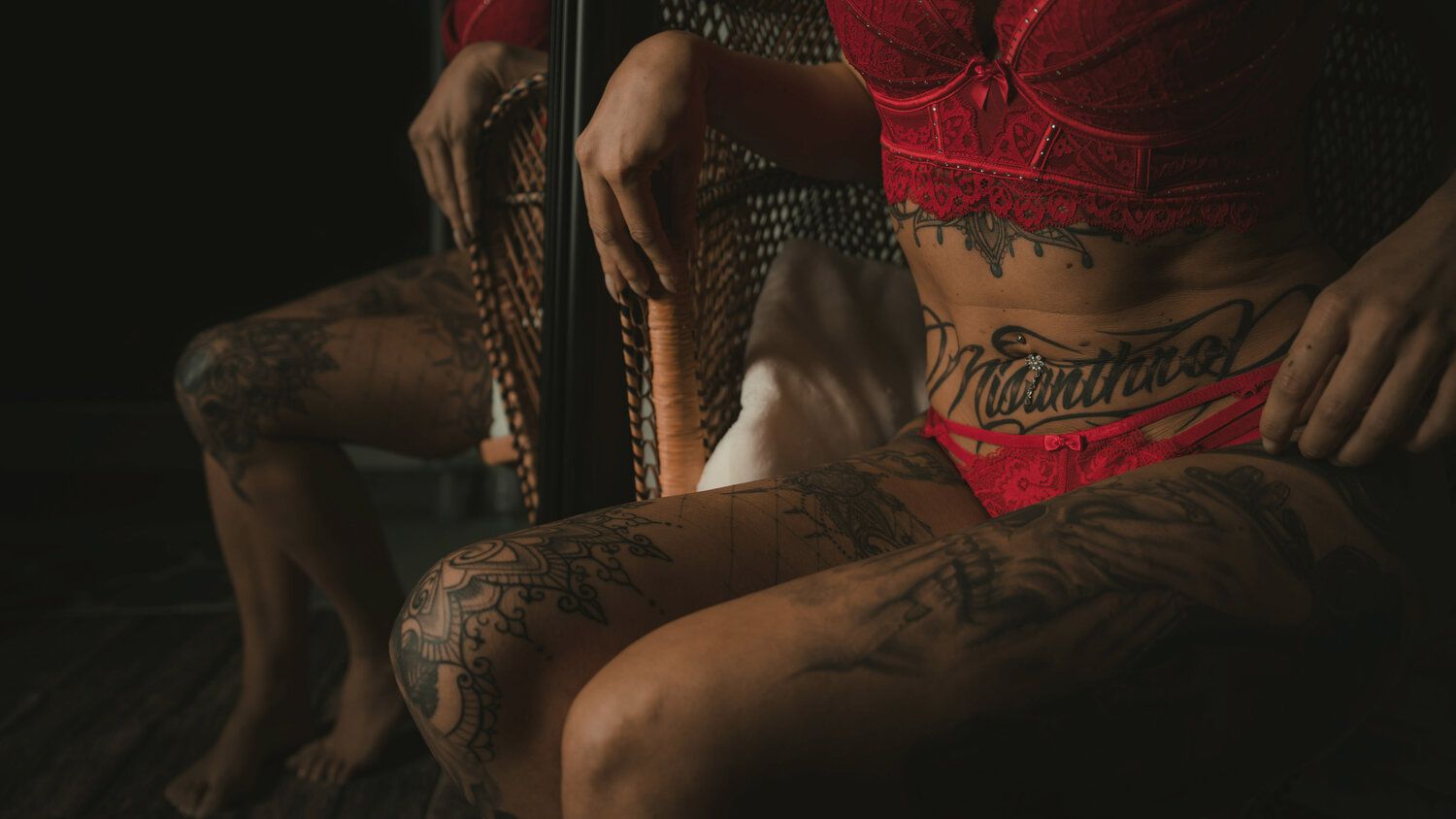 Tattooed person in red lingerie sitting on a wicker chair in dramatic dark lighting.