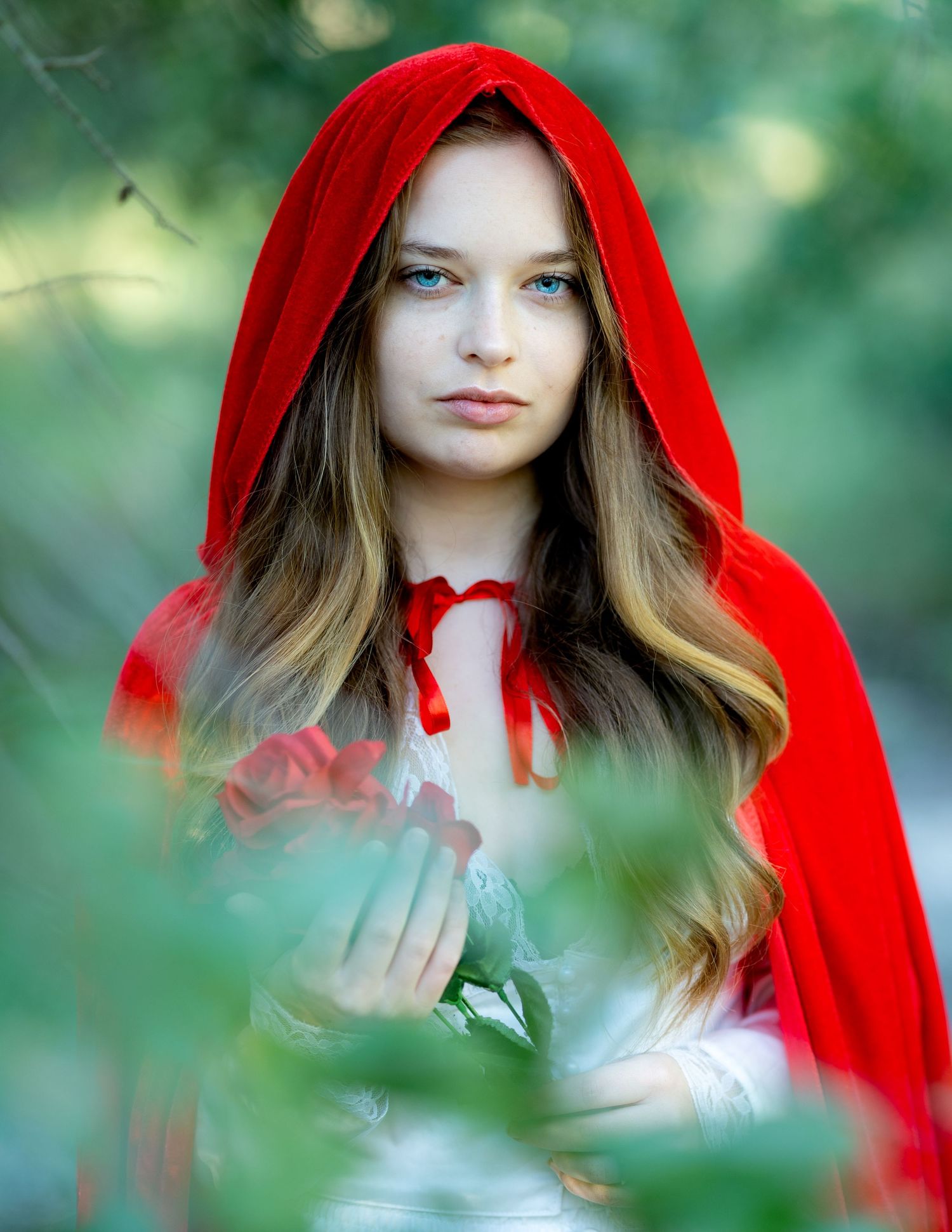 Young woman dressed as Little Red Riding Hood in red velvet cloak, holding roses in a lush green forest setting.
