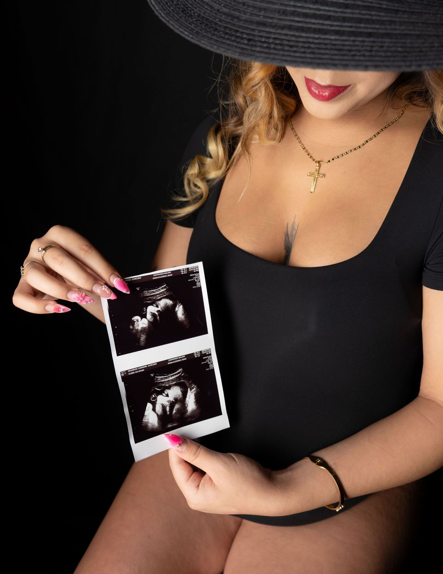 Pregnant woman in black swimsuit and hat holding ultrasound photos, wearing gold cross necklace and pink nails.