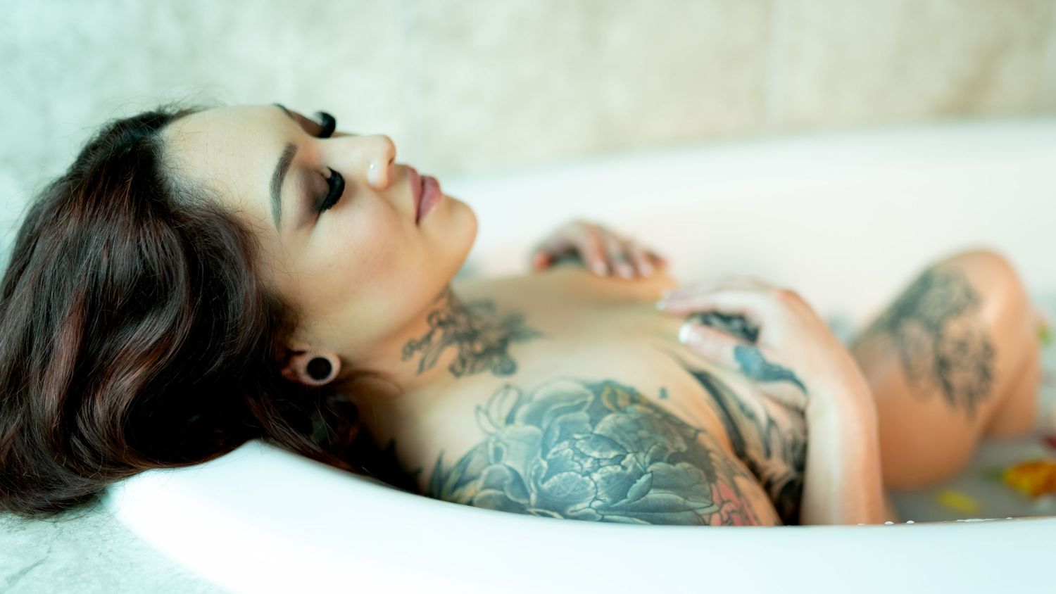 Tattooed woman relaxing in a white bathtub, showcasing intricate body art across her arms and torso.