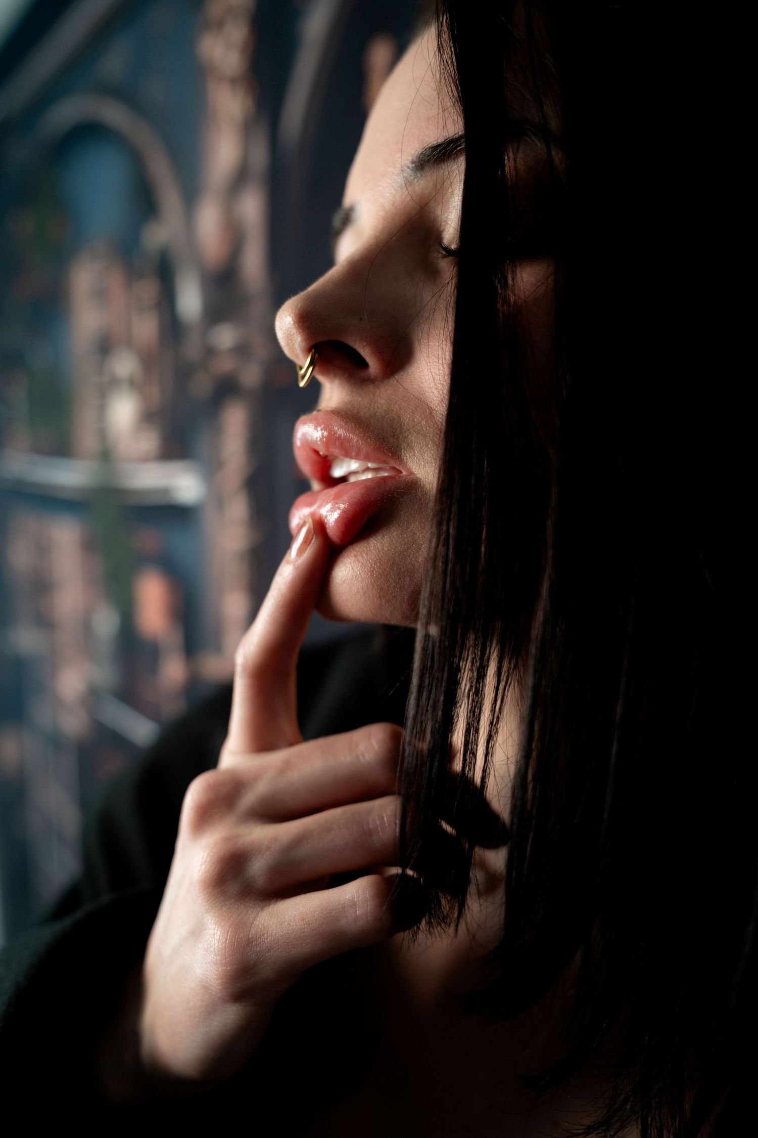 Close-up profile of a dark-haired woman with a nose ring, finger touching her lips in a thoughtful pose.