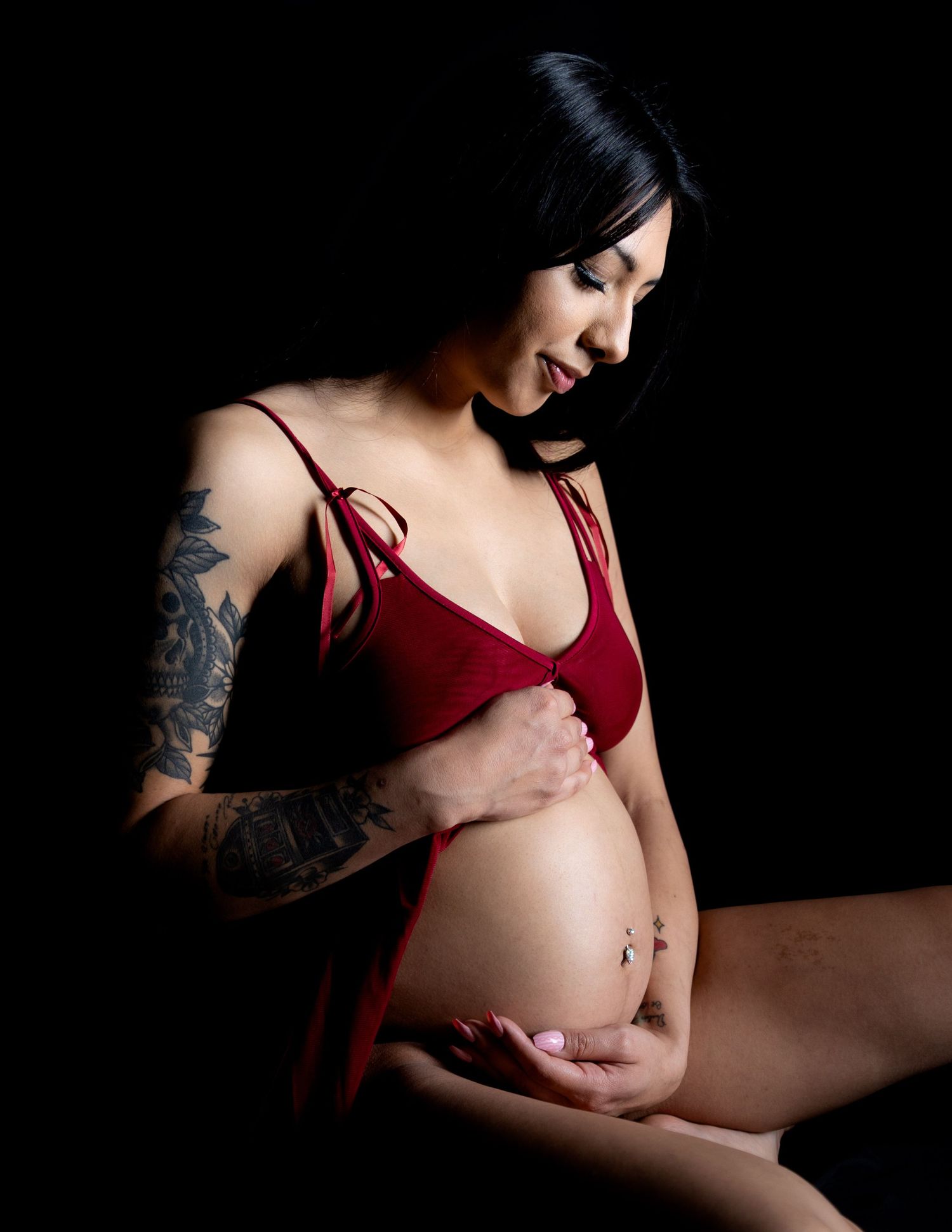 Artistic maternity photos in burgundy lingerie showing pregnant belly against dark background with dramatic shadows.
