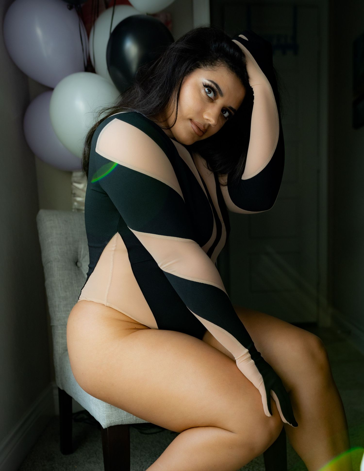 Woman in black and nude bodysuit posing on chair with balloons in background.