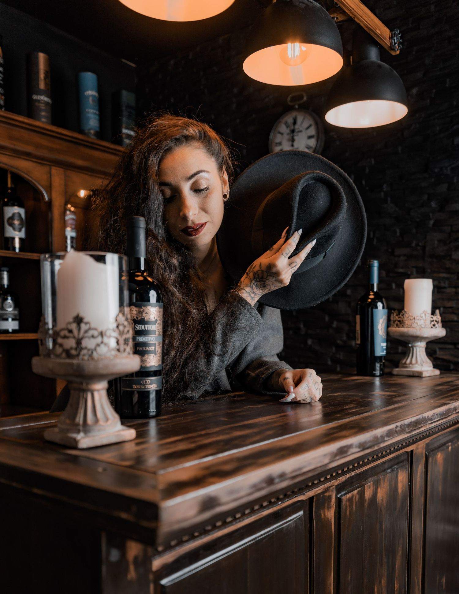 Woman holding black hat at rustic bar with candles, wine bottles, and warm pendant lights in moody setting.