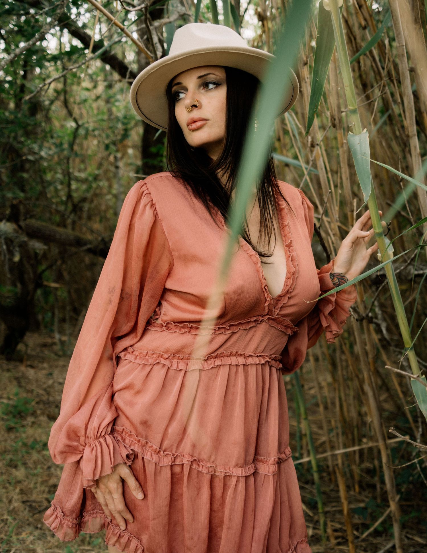 Woman in pink boho dress and wide-brim hat poses among bamboo plants in a natural outdoor setting.