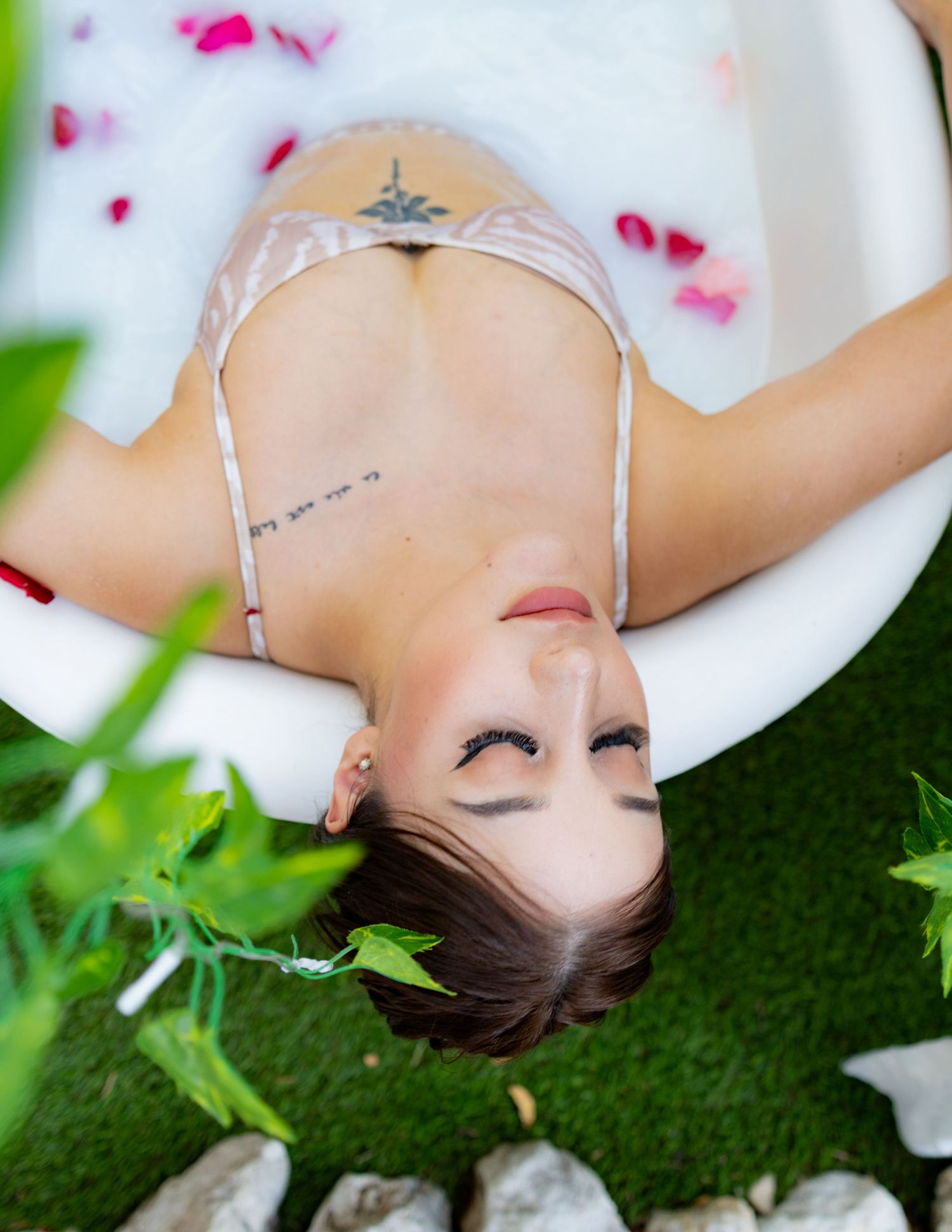 Woman with tattoos relaxing in a floral milk bath outdoors, surrounded by lush green plants and rose petals.