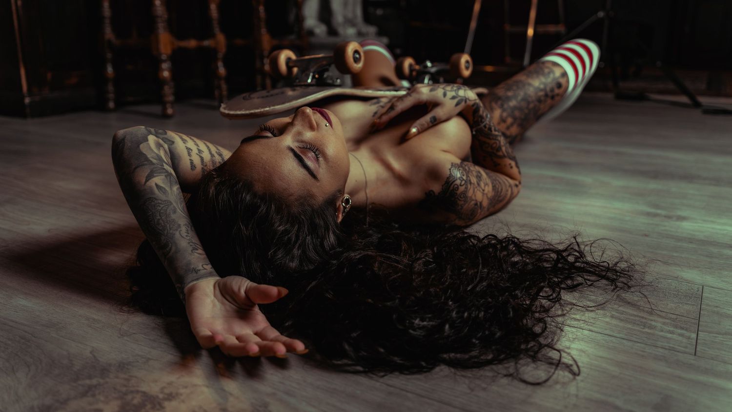 Tattooed woman lying on wooden floor with skateboard, long dark hair spread out, wearing striped socks.