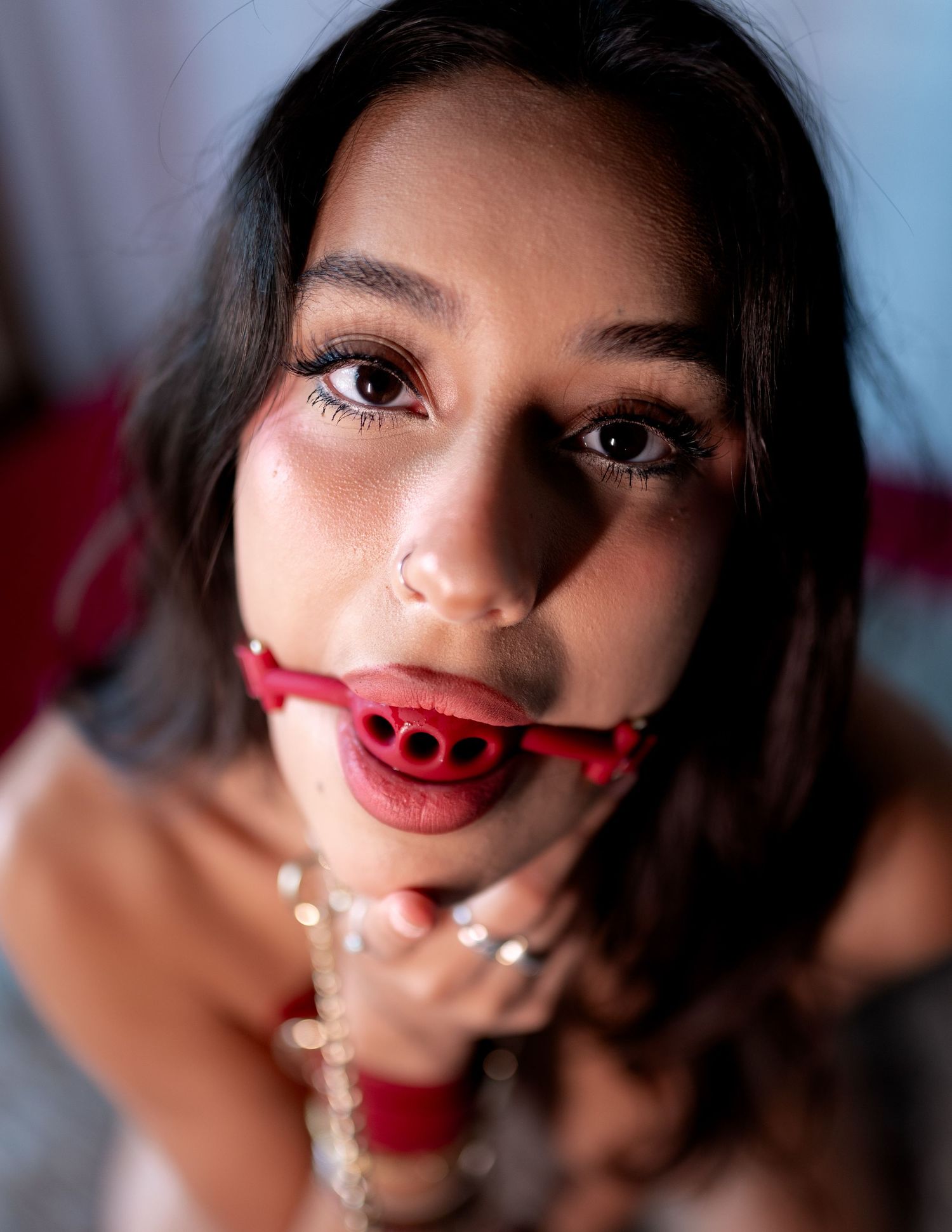 Young woman with dark hair wearing a red ball gag and chain necklace, looking up at camera.